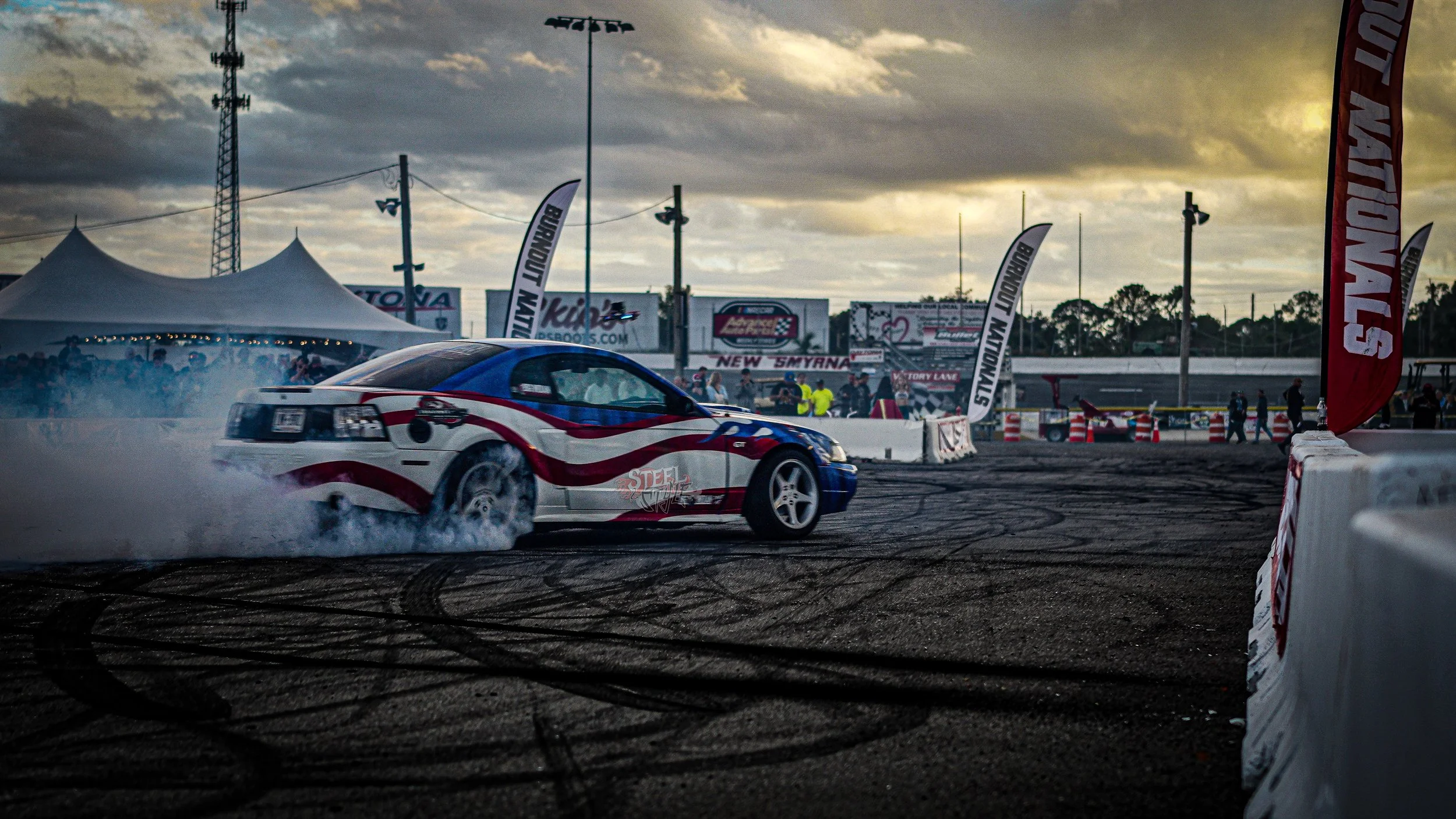 A race car performing a burnout on a racetrack at sunset, with smoke billowing from the tires, surrounded by race banners and spectators in the background.