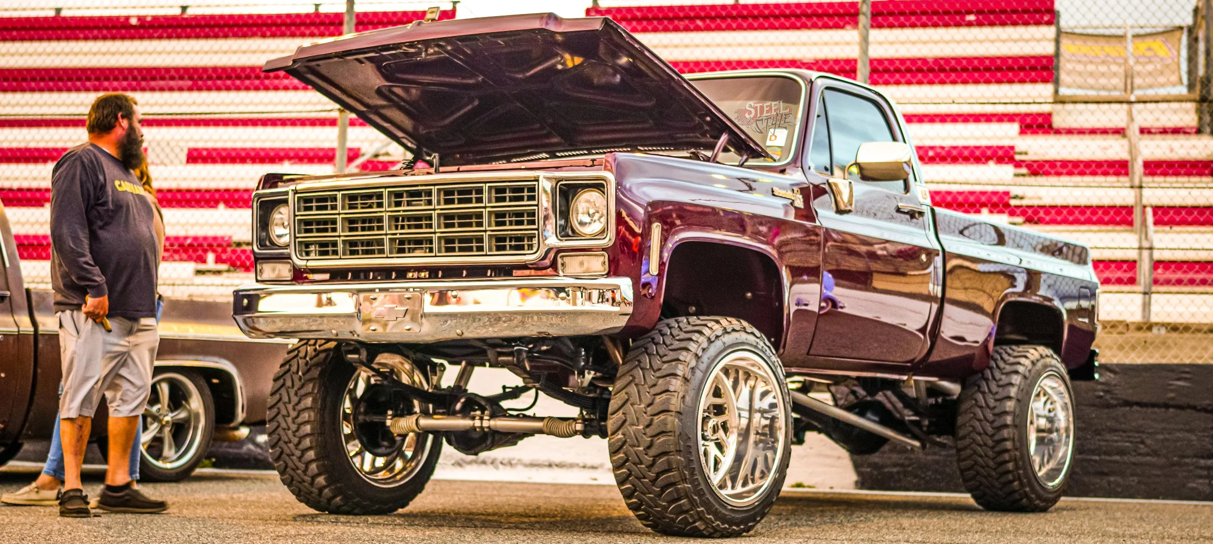 A lifted maroon Chevrolet pickup truck at a car show, with the hood open and large off-road tires, parked near bleachers with red and white seats, and a man with a beard wearing a dark long-sleeve shirt and shorts standing next to it.