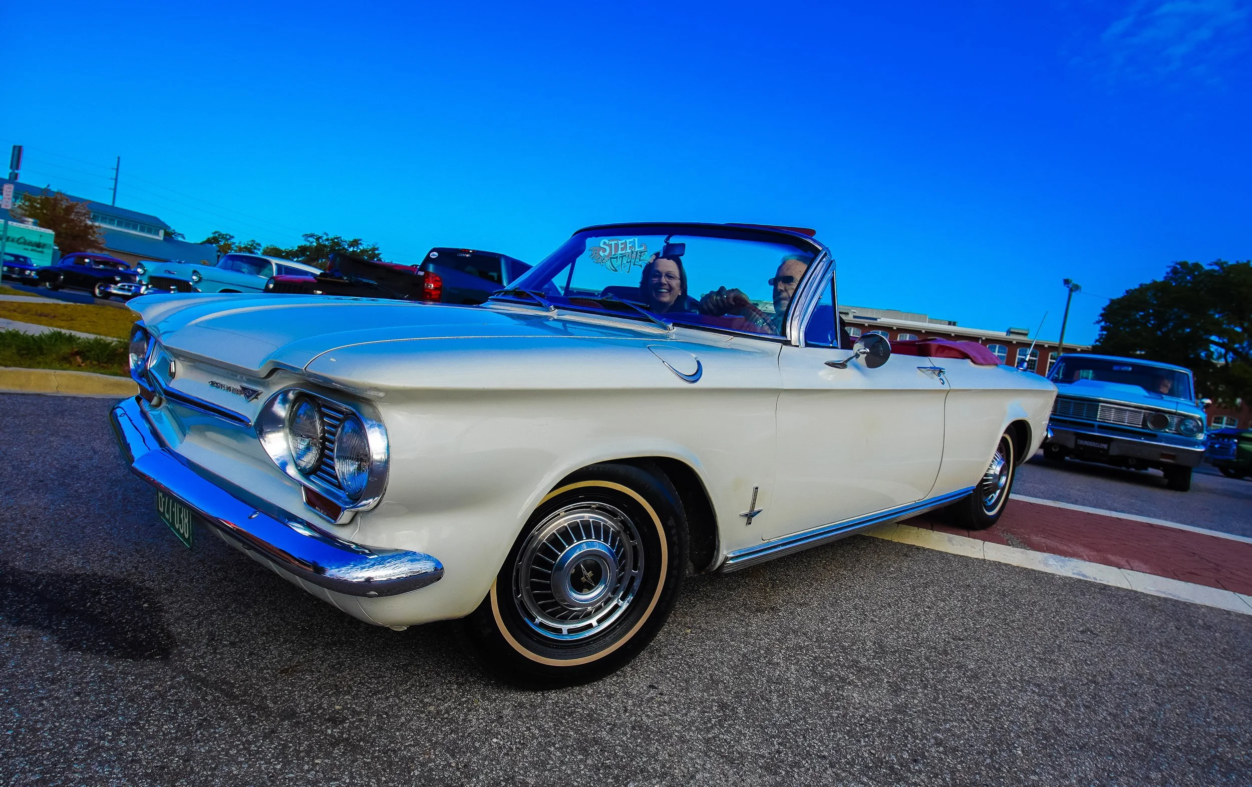 A white vintage convertible car parked at a classic car show with two smiling people inside, one man driving and one woman in the passenger seat, on a sunny day with a clear blue sky.