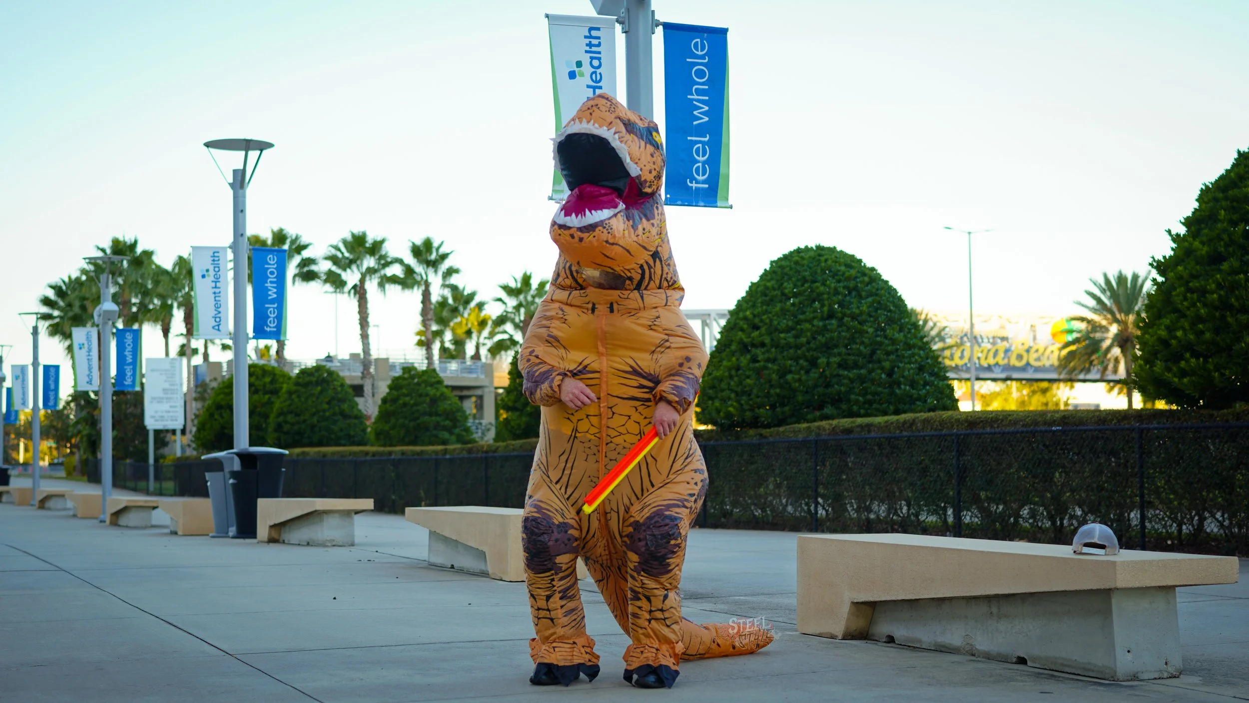 Person dressed in a complete T. rex dinosaur costume standing on a sidewalk at an outdoor location, with palm trees and signage in the background.
