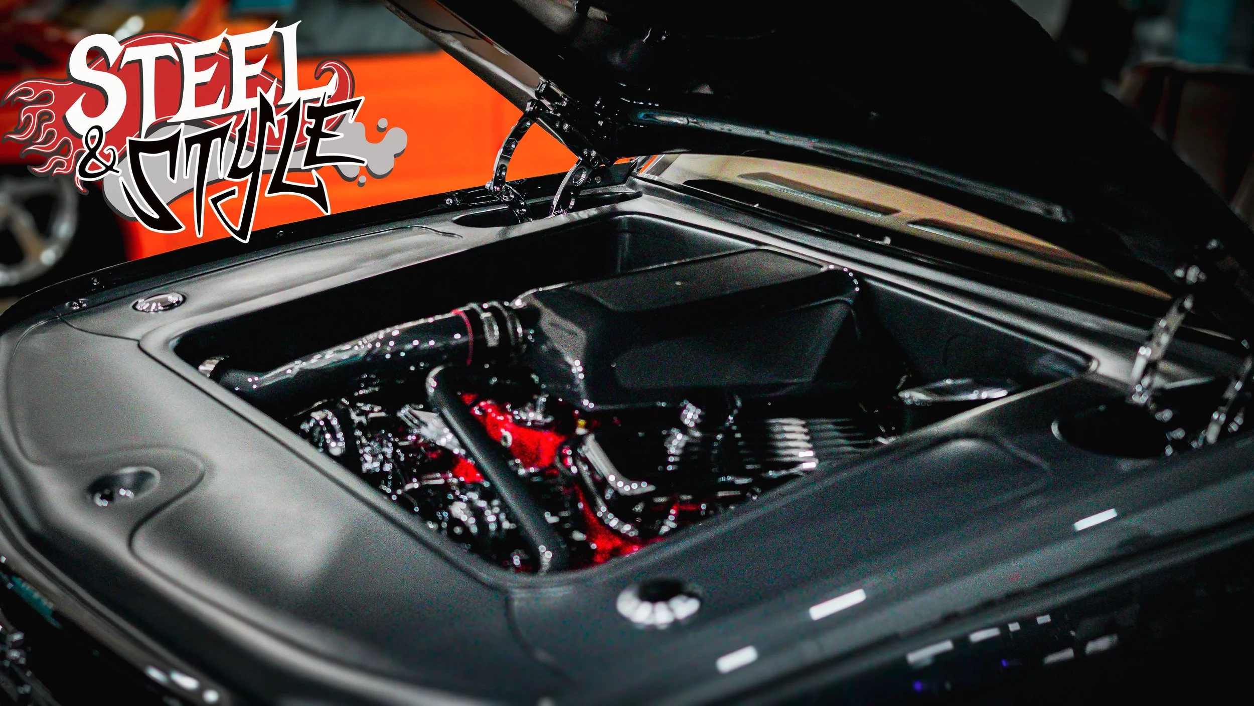 Close-up of a car engine with the hood open, showing various engine parts in black and red, with a graffiti-style logo reading 'Steel & Style' in the background.