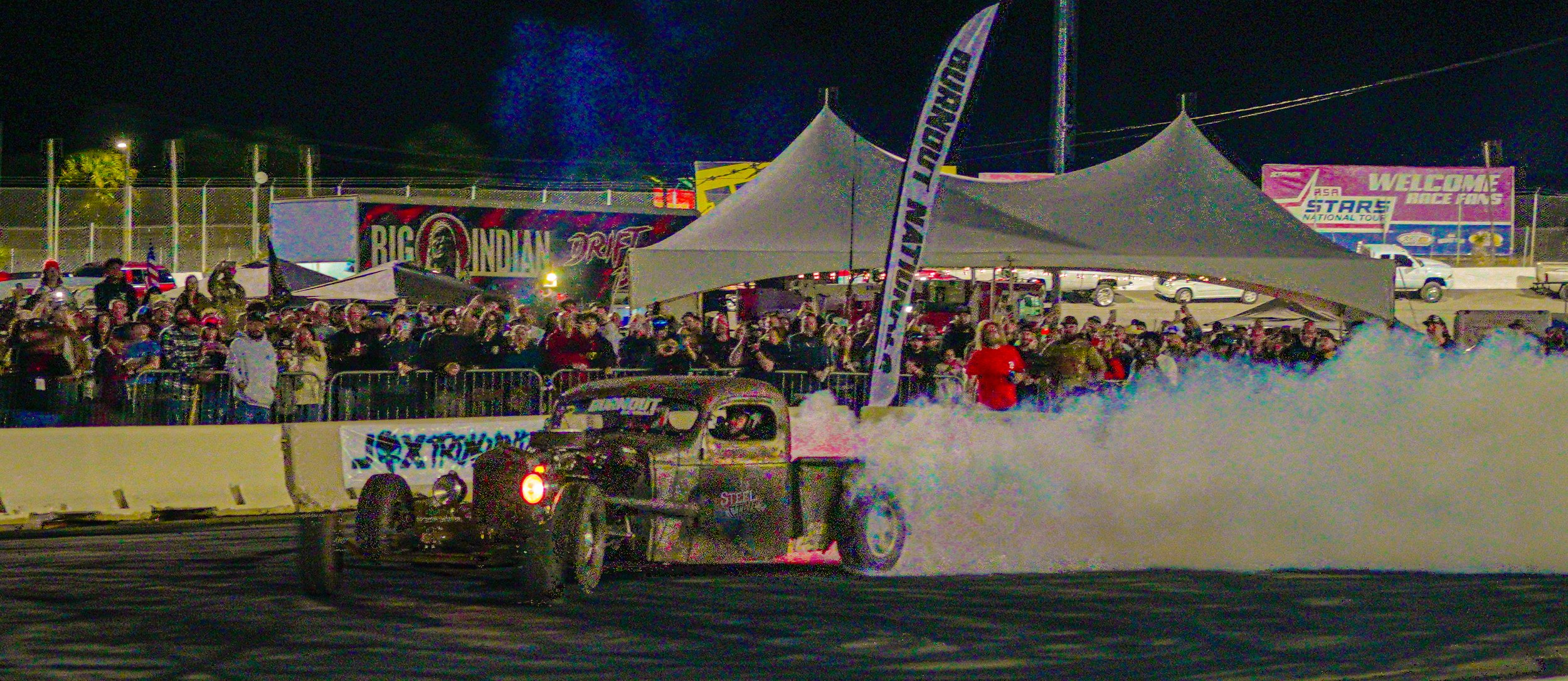 Nighttime race event with a vintage car drifting, kicking up smoke on the track, crowd watching behind barriers, tents, and banners in the background.