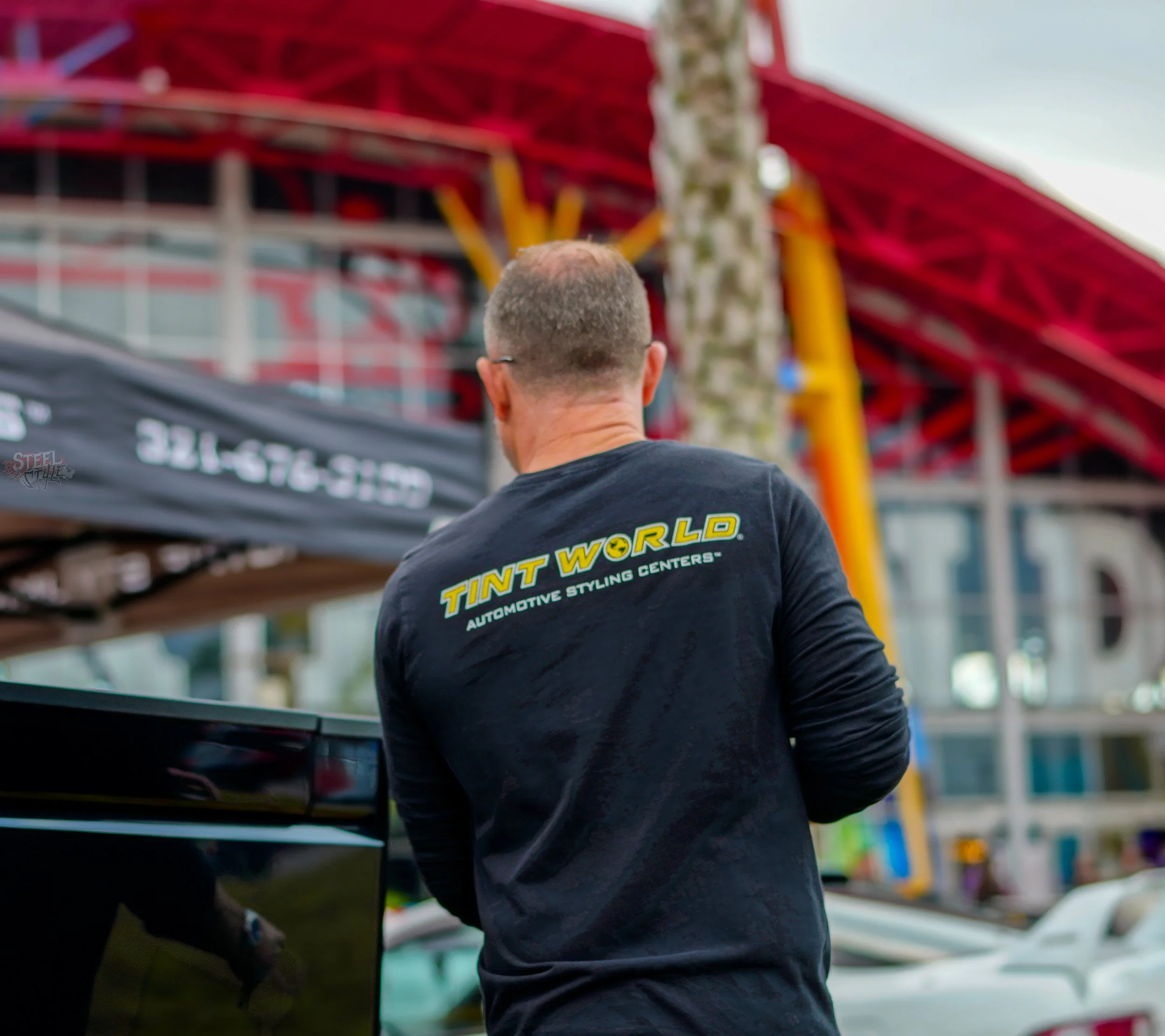 A man with short hair wearing a black long-sleeve shirt with yellow text that says 'TINT WORLD' and 'AUTOMOTIVE STYLING CENTERS' on the back, standing outdoors with his back to the camera, near a black vehicle and a red-framed structure in the backgr