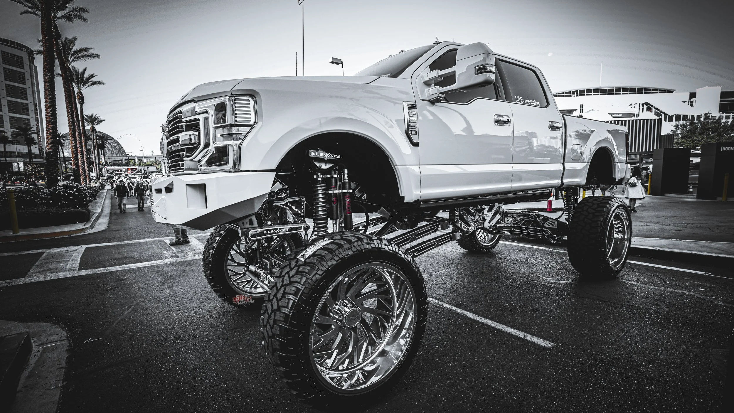 White lifted truck with oversized black tires on display outdoors in an urban area with palm trees and pedestrians in the background.