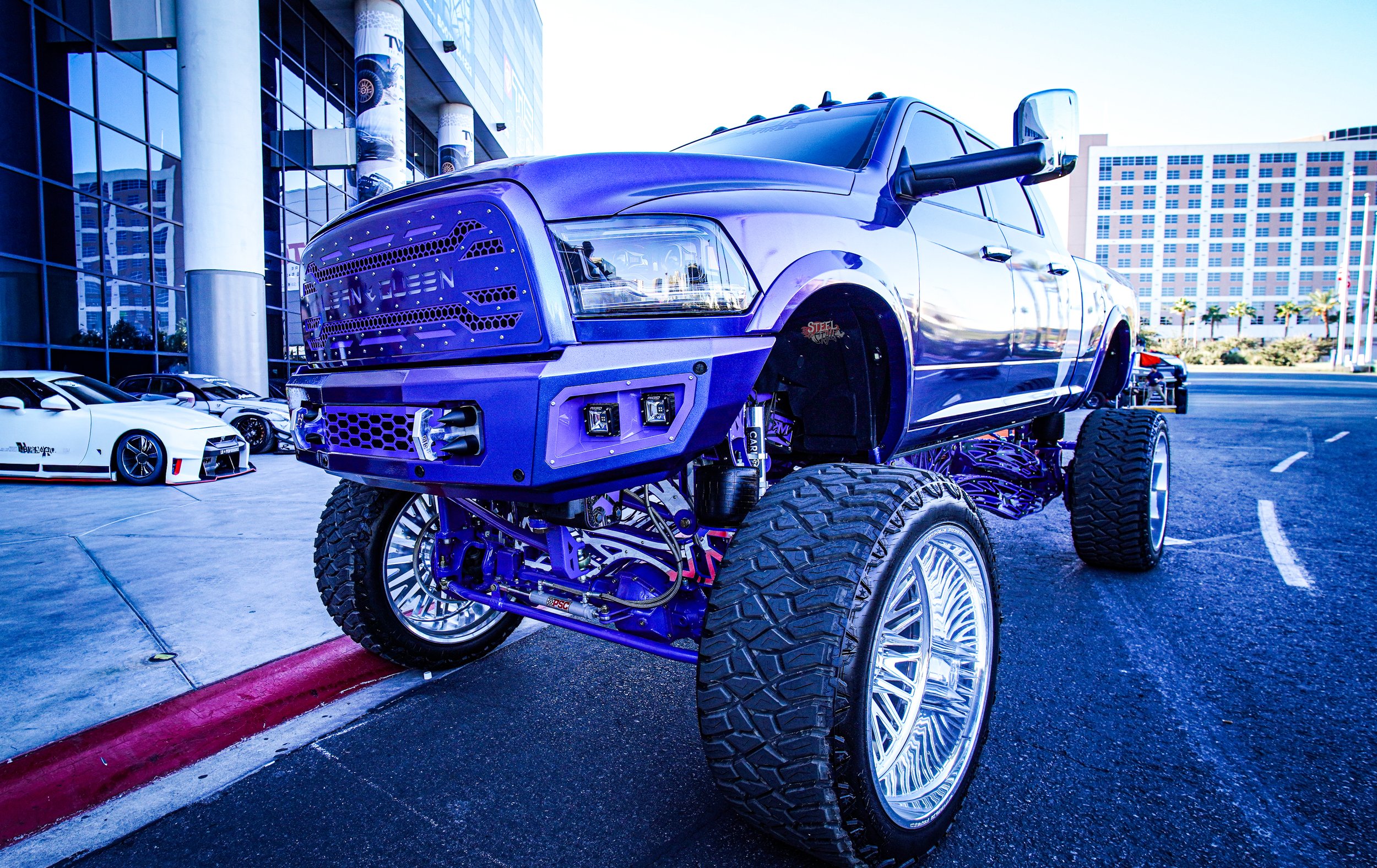 A modified pickup truck with oversized wheels, painted purple, parked outside a modern building with reflective glass windows in an urban area, with sports cars parked nearby.