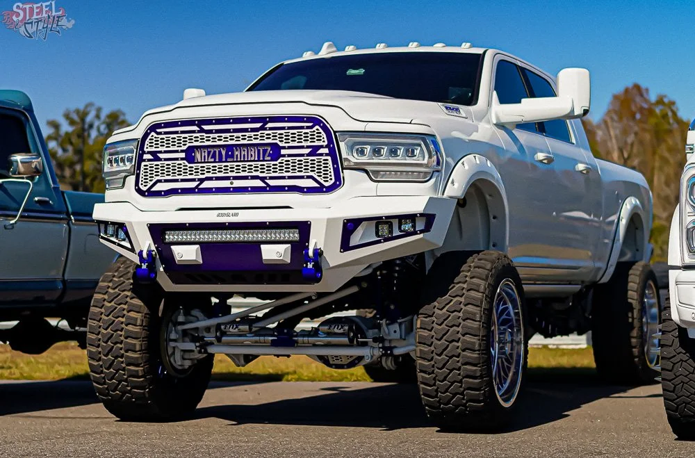 A white lifted pickup truck with oversized tires, custom front bumper, and a license plate that reads 'MIZTY-HARITZ,' parked outdoors on pavement with trees and blue sky in the background.