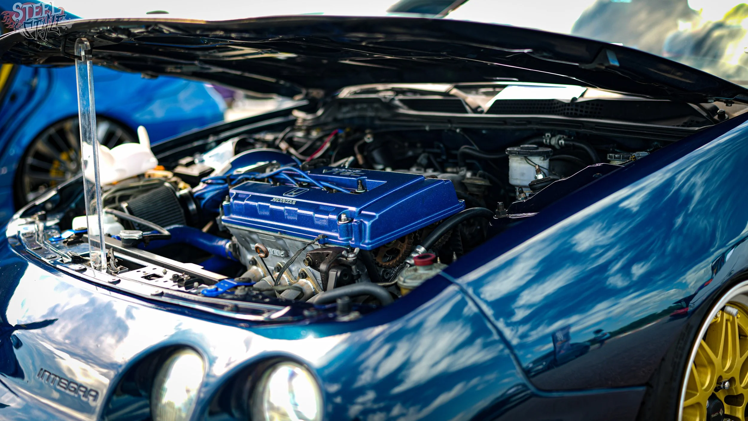 Close-up of a car engine inside an open hood, with a blue valve cover and yellow wheels visible on the car.