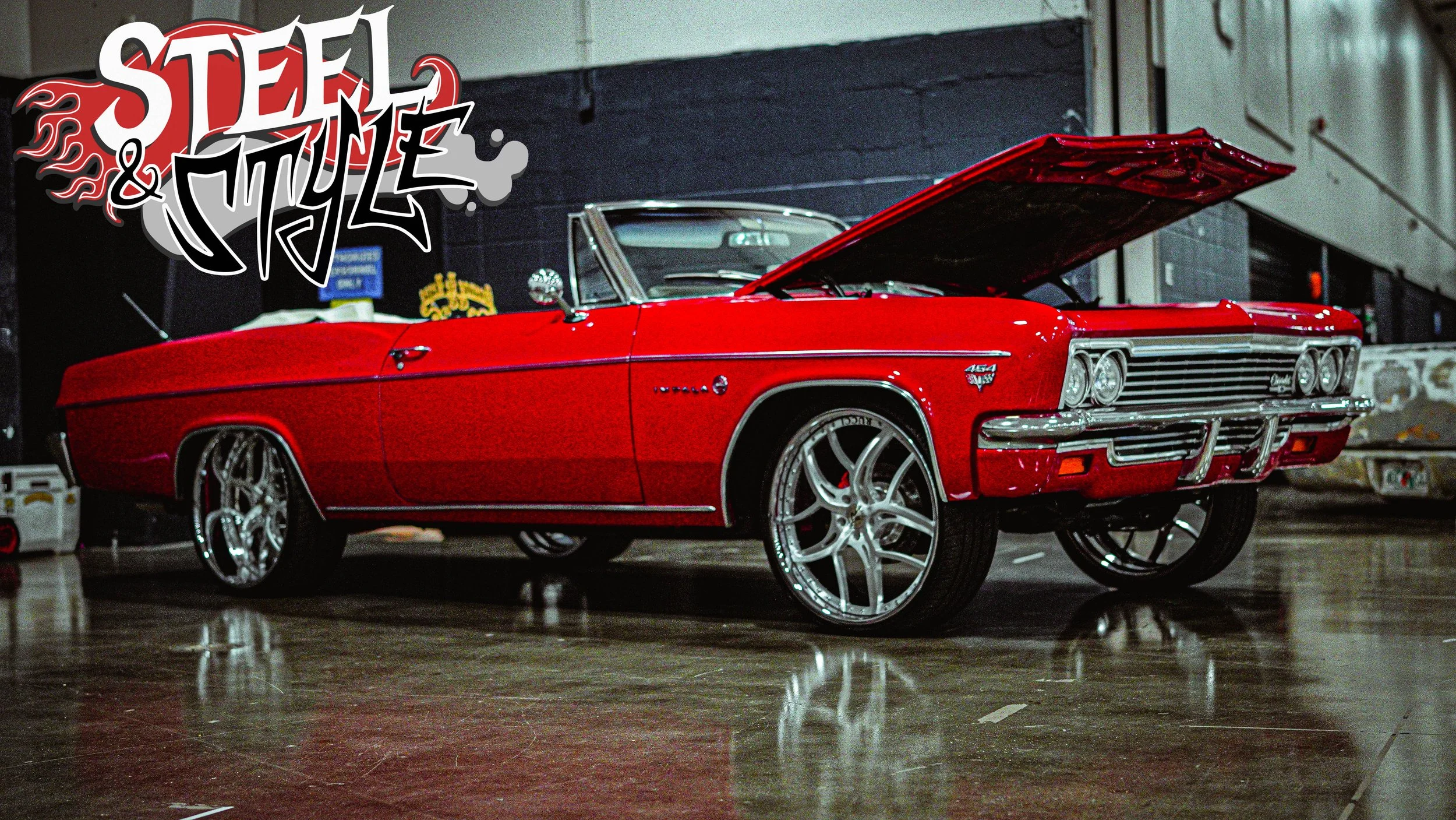 Red vintage convertible car with its hood open, parked indoors, with custom rims and chrome detailing, in a garage or showroom setting.