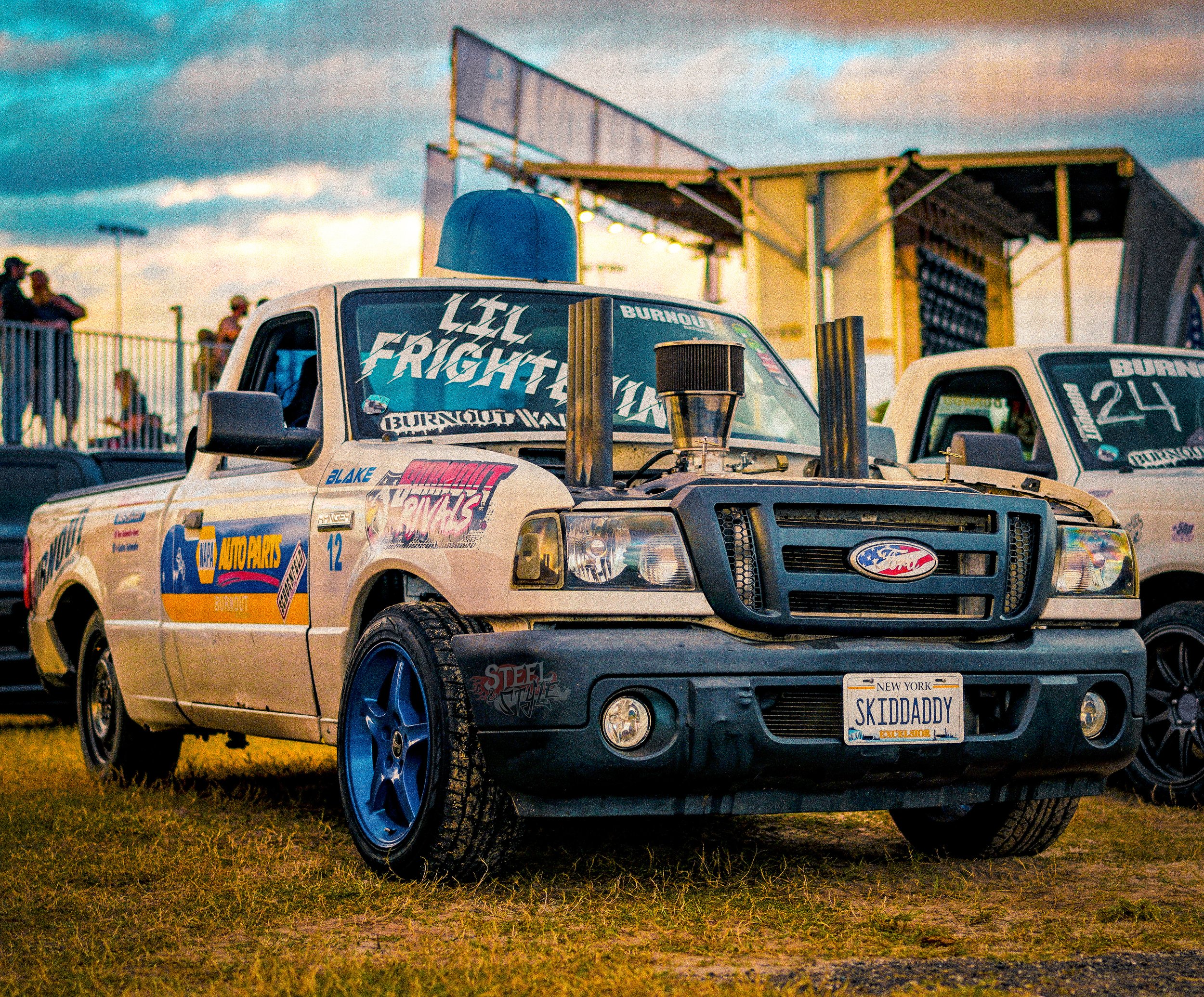A modified Ford pickup truck with racing decals and a custom license plate reading 'SKIDDADDY' parked on grass at a race track during sunset. Other similar trucks and spectators are visible in the background.