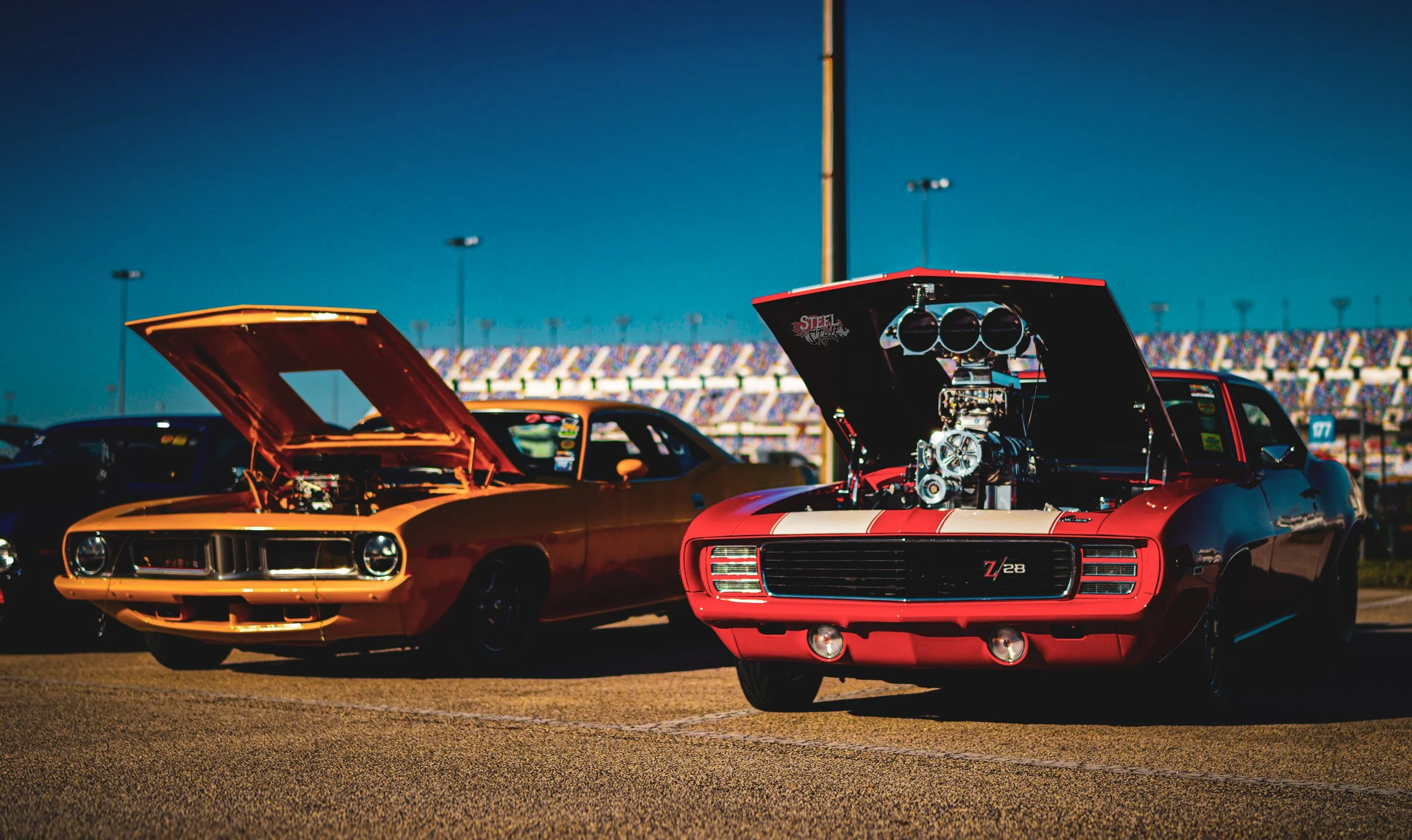 Two classic muscle cars parked on a racetrack with their hoods open, exposing their engines during a sunny day with a clear sky.