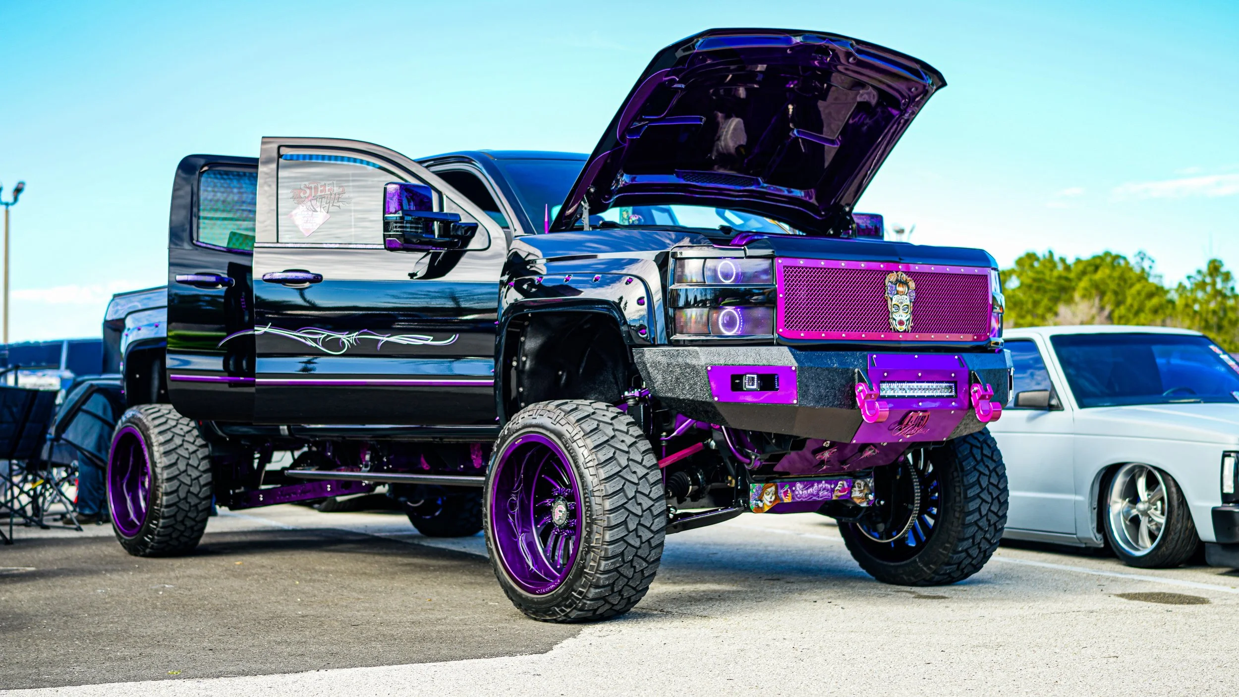 A customized lifted pickup truck painted in black with purple accents and chrome detailing, parked outdoors with its hood open and next to a white vehicle.