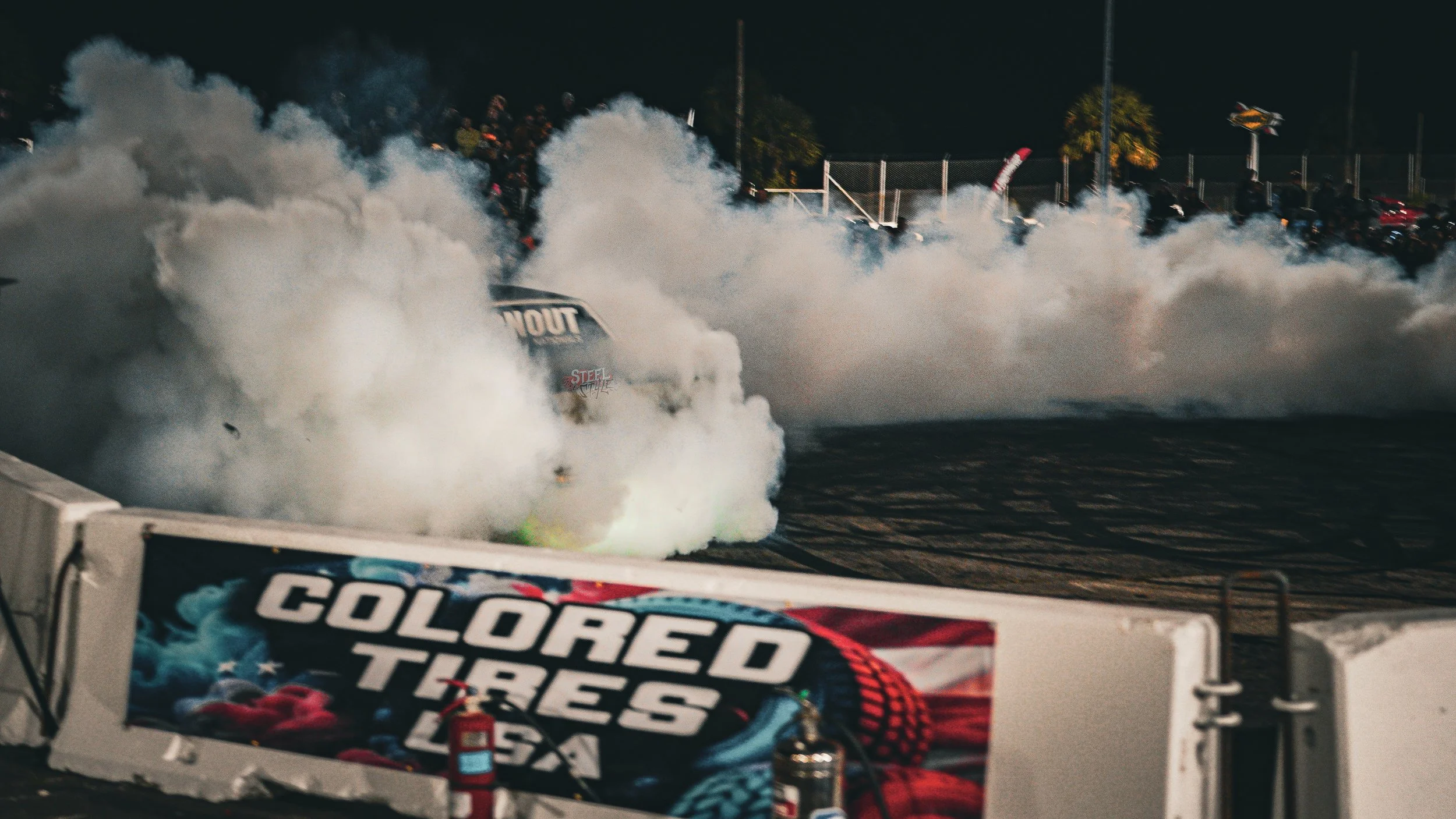A race car spins out on the track, creating a large cloud of smoke. A banner in the foreground reads 'Colored Tires USA' and there are spectators and equipment visible in the background.