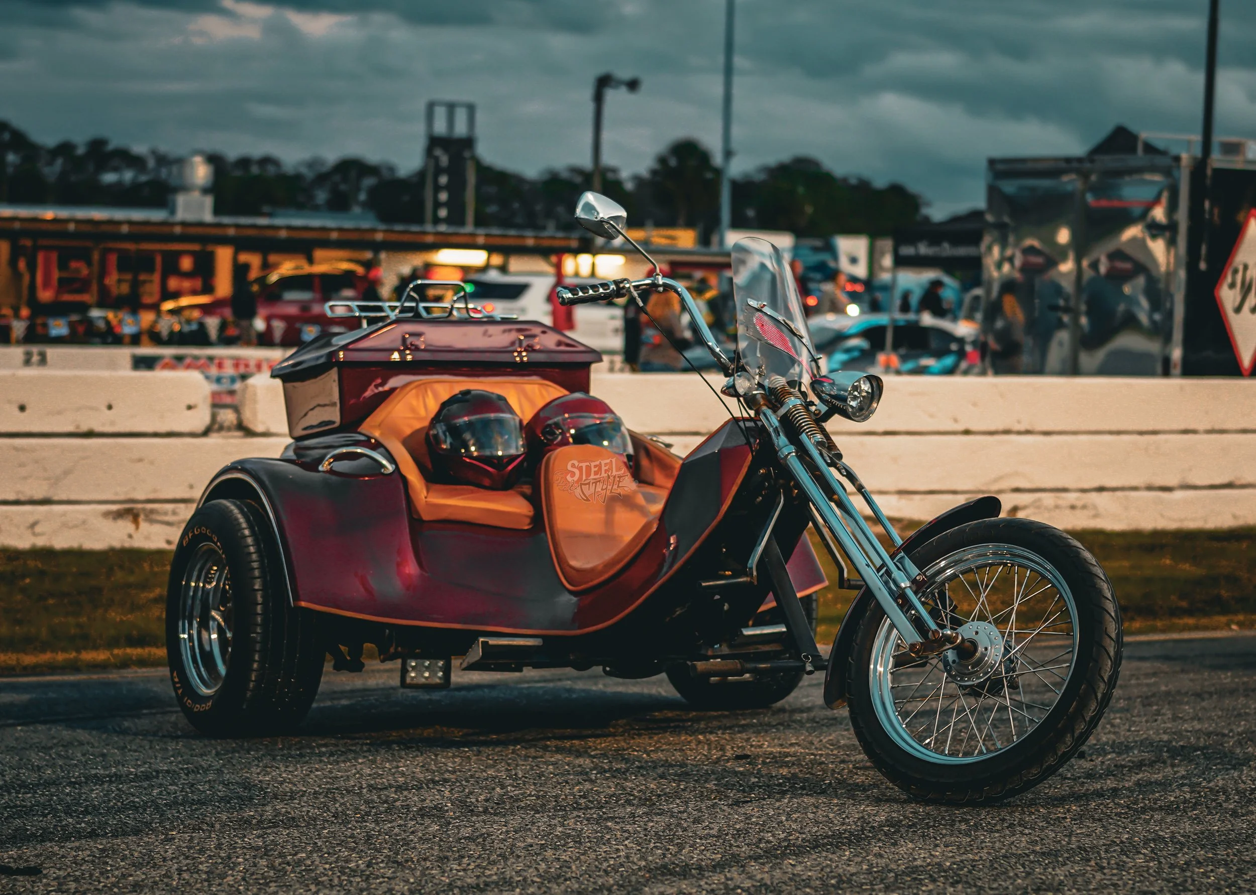 A red and black motorcycle with a sidecar, parked outdoors during twilight with a busy background of cars and people.