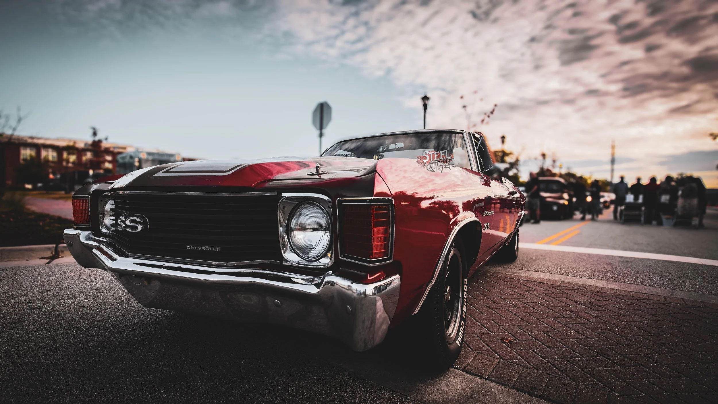 Red Chevrolet muscle car parked on a city street during sunset with a group of people in the background.