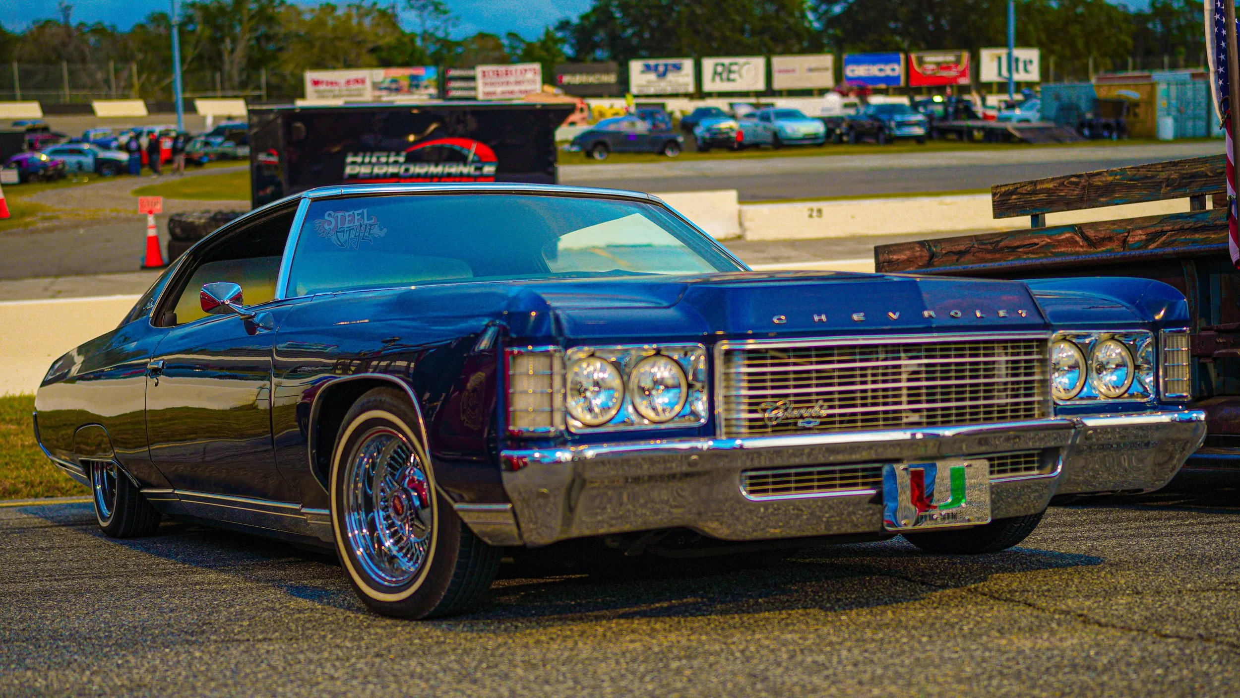 A classic blue Chevrolet car parked at a car show, with other vintage vehicles and a parking lot in the background.