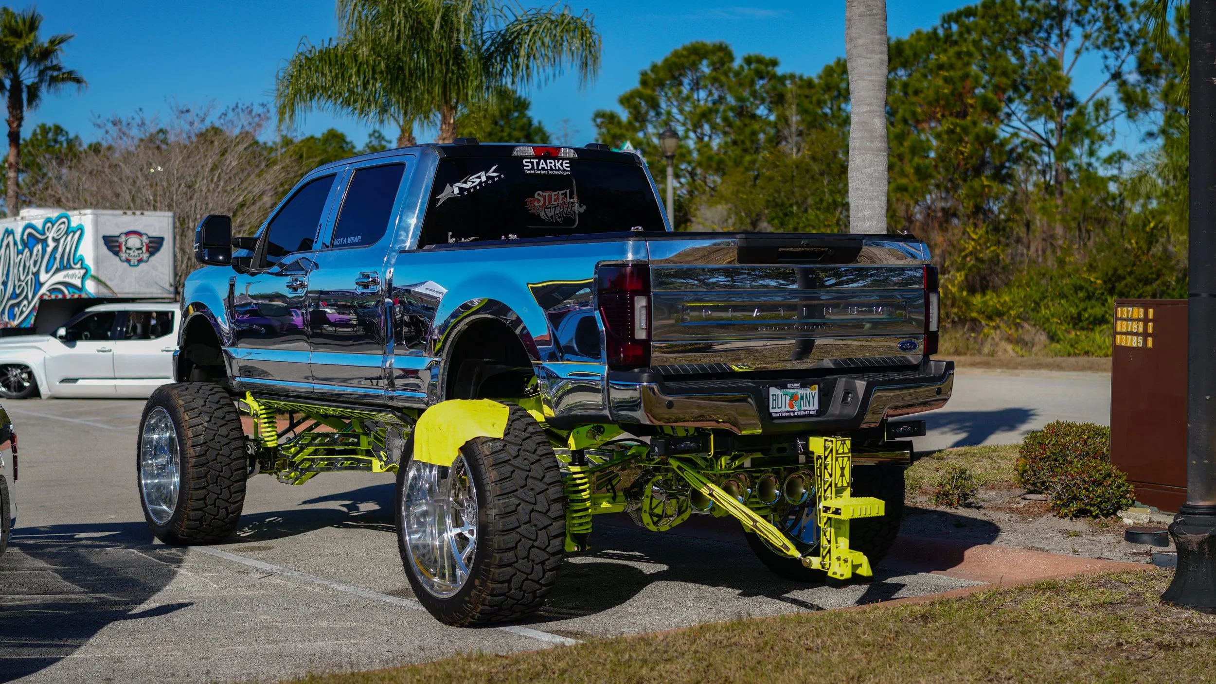 A customized blue Ford pickup truck with a chrome finish and a yellow suspension lift, parked in a lot on a sunny day with palm trees and other vehicles in the background.