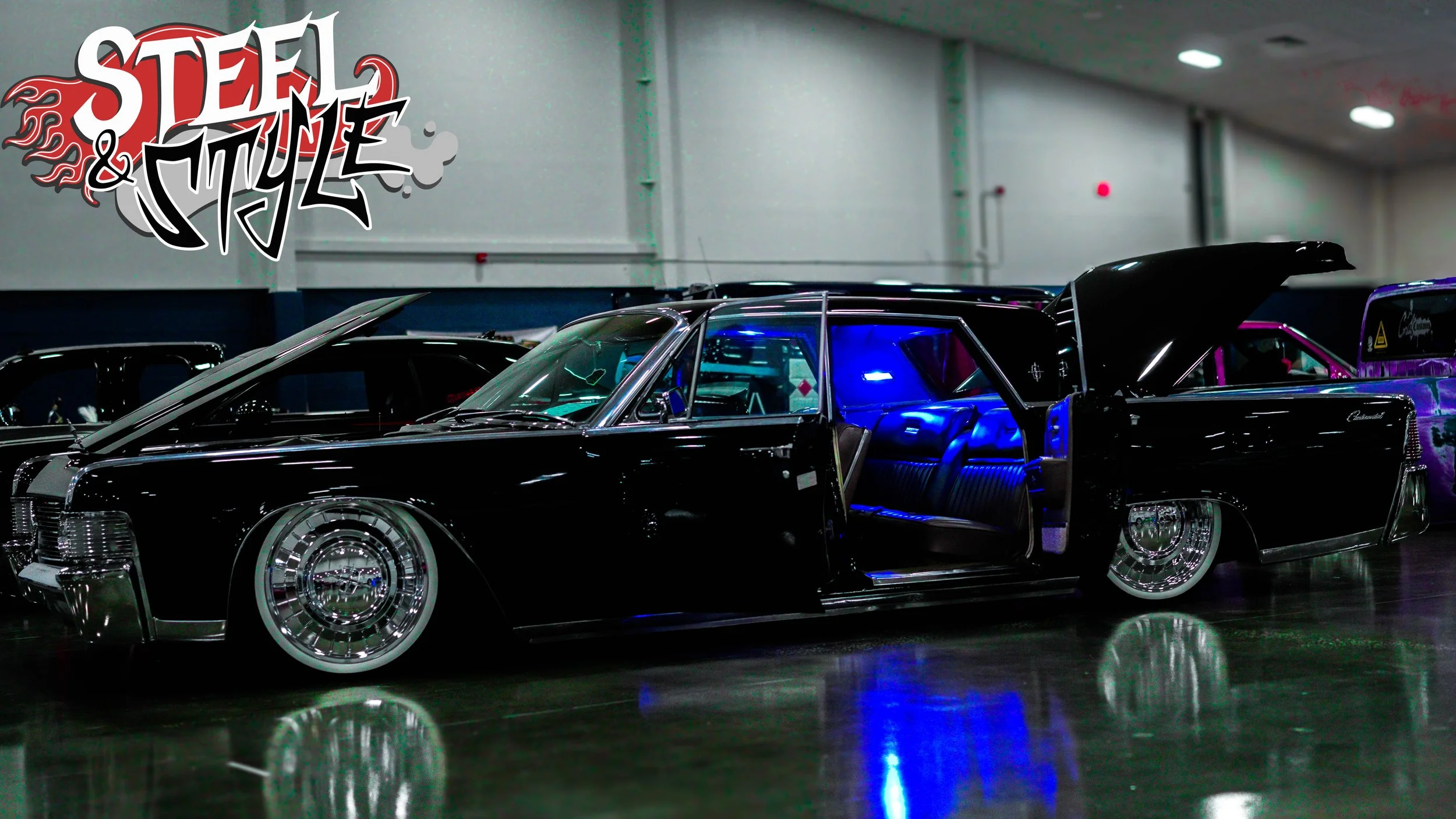 A vintage black Cadillac with its doors and trunk open, parked indoors during a car show. The interior is illuminated with blue lighting, and other classic cars are visible in the background.