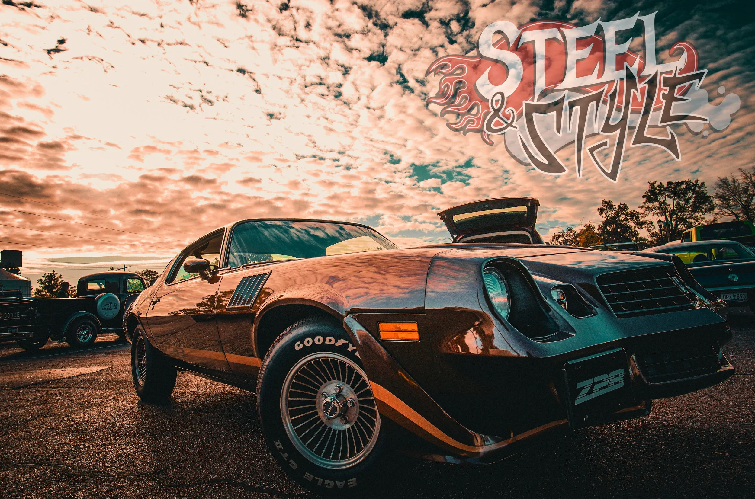 A classic black Pontiac Firebird Trans Am with gold accents parked outdoors during sunset, with vintage cars in the background and clouds in the sky, featuring a 'Steel & Mettle' logo in the top right corner.