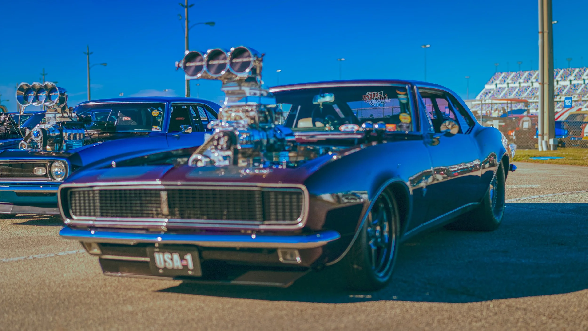 A black vintage muscle car with a large supercharger on the hood at a racing event, parked on asphalt with a blue sky background. Another muscle car with a similar supercharger is partially visible behind it.