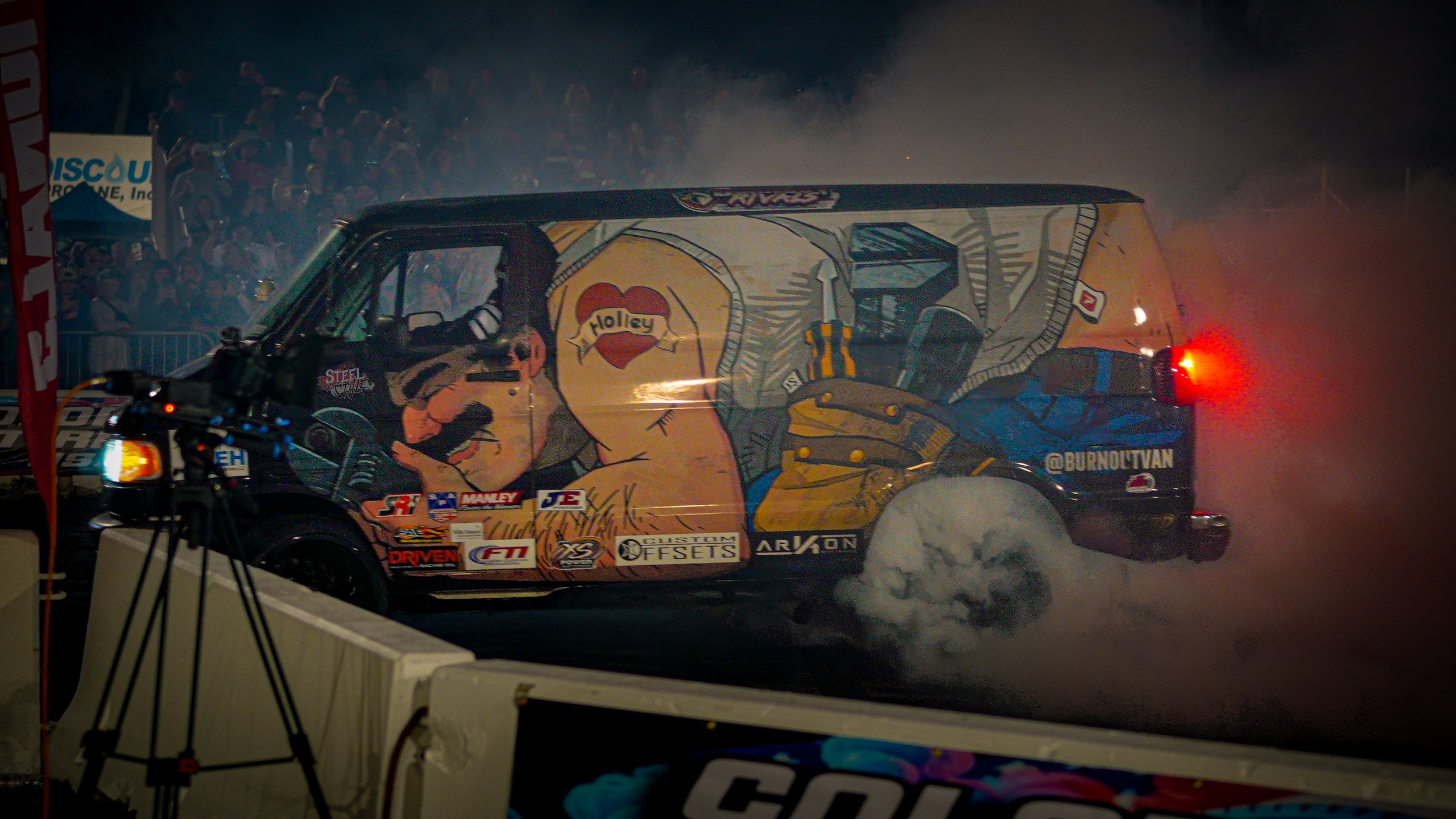 A race car performing a burnout at night with smoke billowing from the tires, featuring colorful artwork of a muscular cartoon character with a heart and the word "Holley" on the side, and a crowd of spectators in the background.