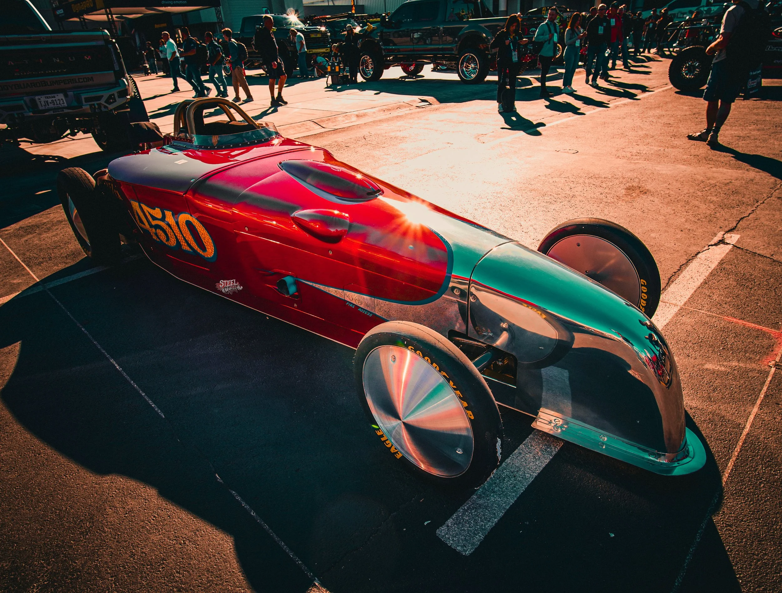 A vintage race car with a chrome body and red, teal, and yellow paint scheme on display at an outdoor car show with people walking around in the background.