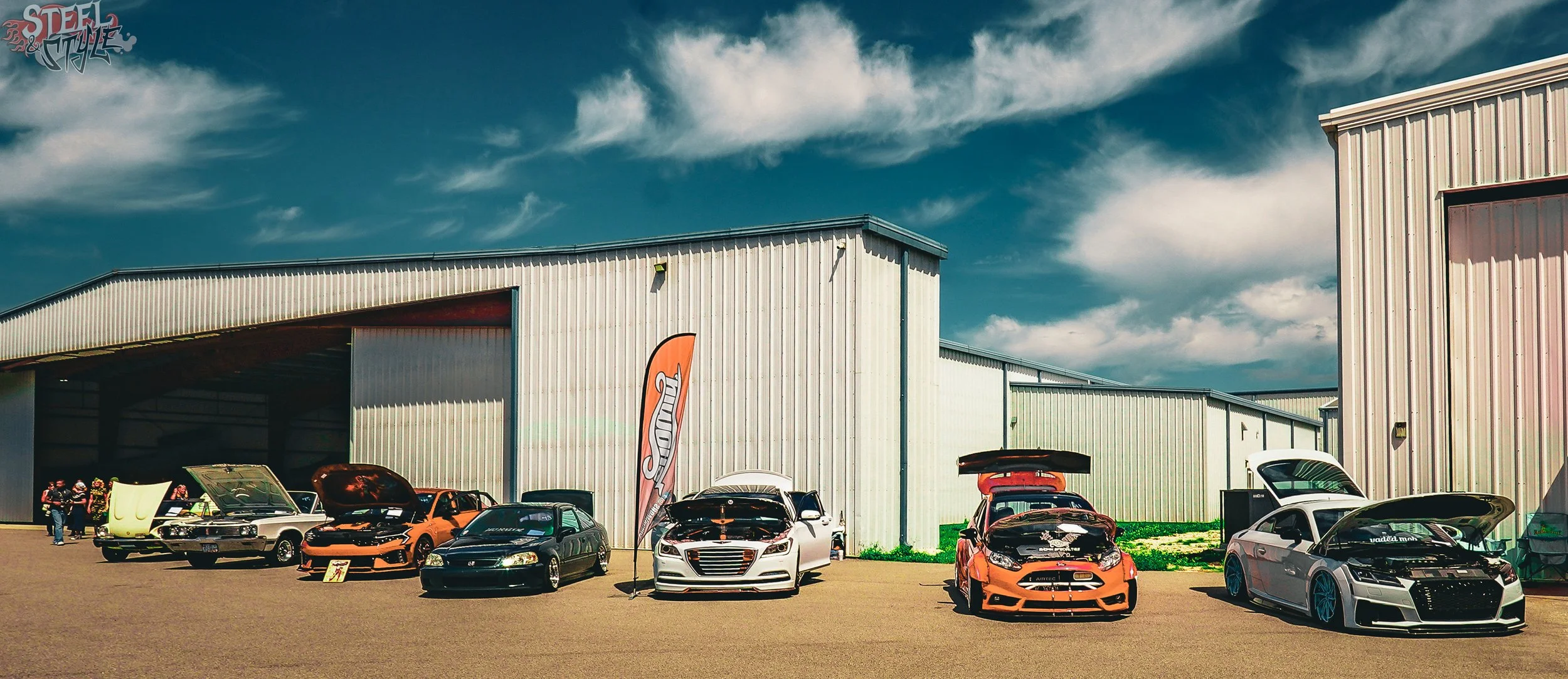 Cars on display at an outdoor car show in front of a large industrial building, with people walking around and a flag featuring the 'Steele & Style' logo.