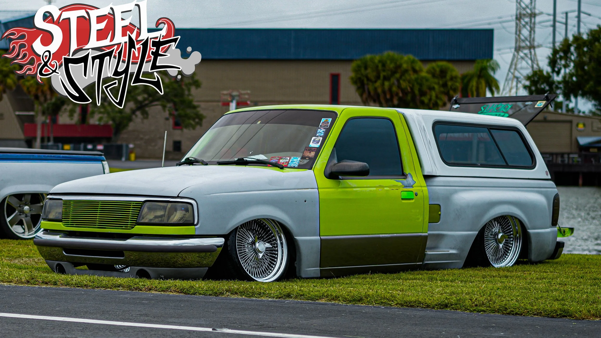 Low-rider vintage car with custom wire wheels and green and silver paint, parked on grass near water with trees and buildings in the background, alongside other vehicles, and a "Steel & Style" logo in the upper left corner.