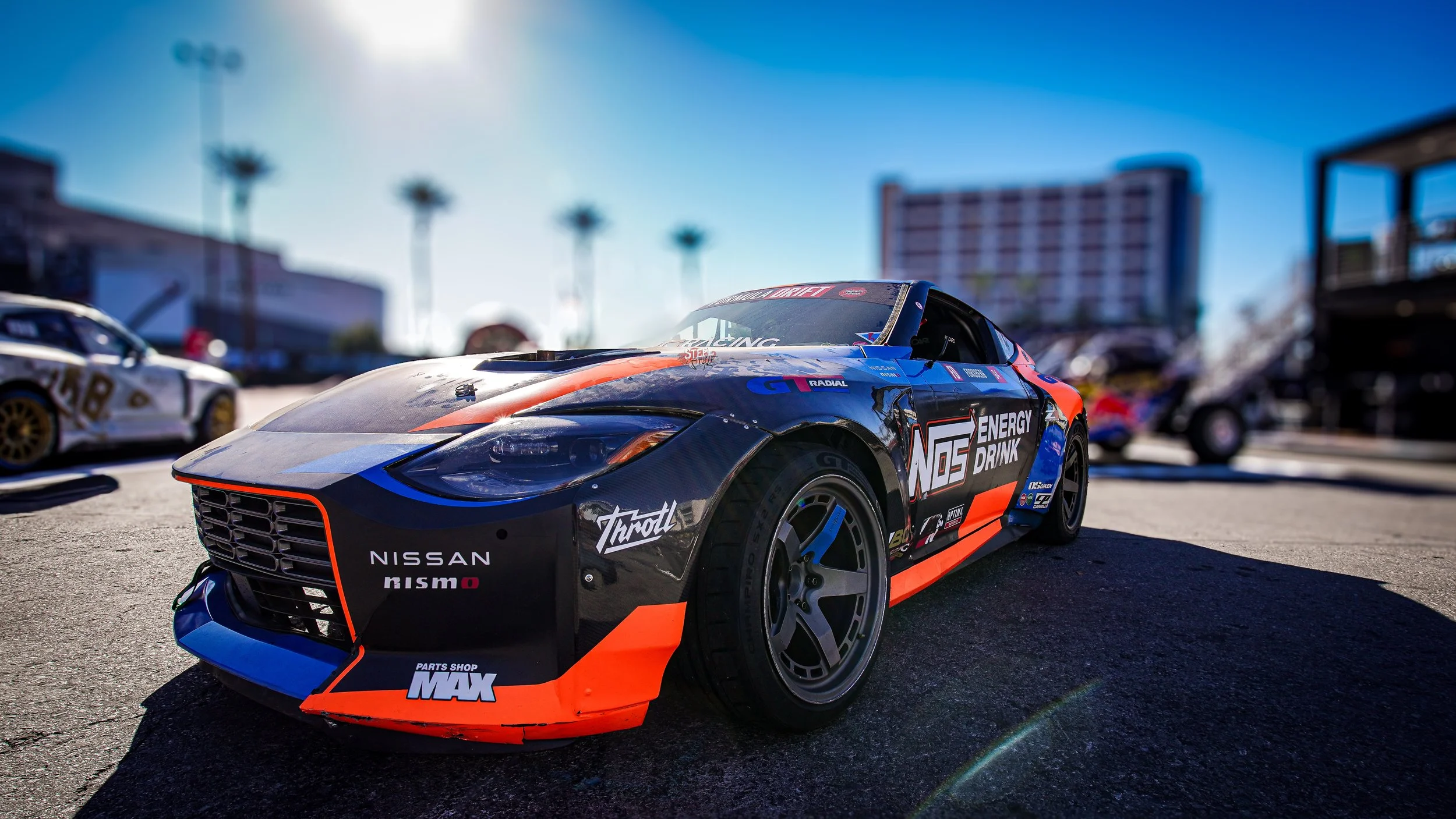 A black race car with orange and blue accents, sponsored by Nissan and Nismo, parked on a paved surface at a race track amid other vehicles with palm trees and buildings in the background, under a bright blue sky.
