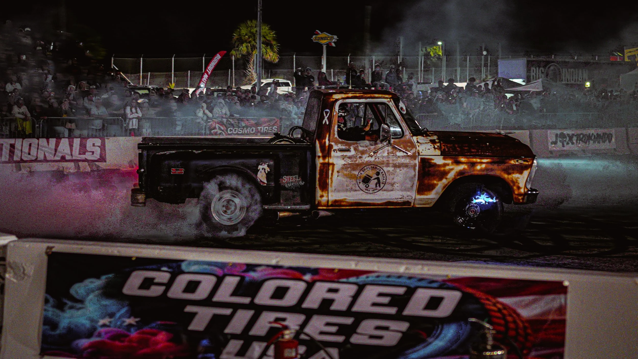 A rusty vintage pickup truck performing a burn-out at night, surrounded by a cloud of smoke and tire smoke, with a crowd of spectators watching behind a fence.