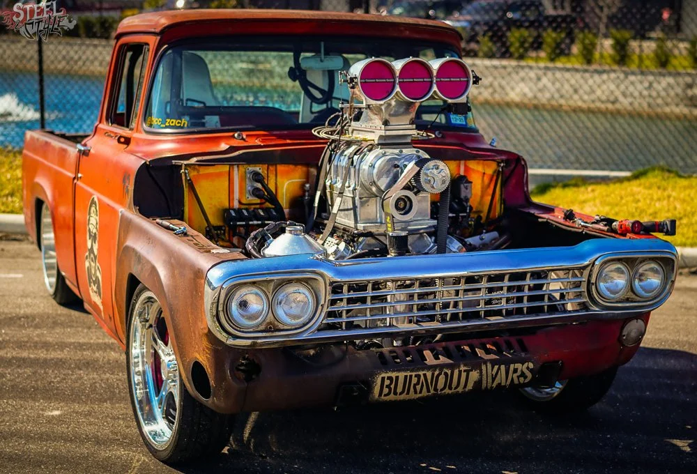 A vintage orange pickup truck with a high-performance engine modification, including a large supercharger with three chrome intake throttles with pink covers, visible under the hood. The truck has racing decals, custom rims, and a license plate that 