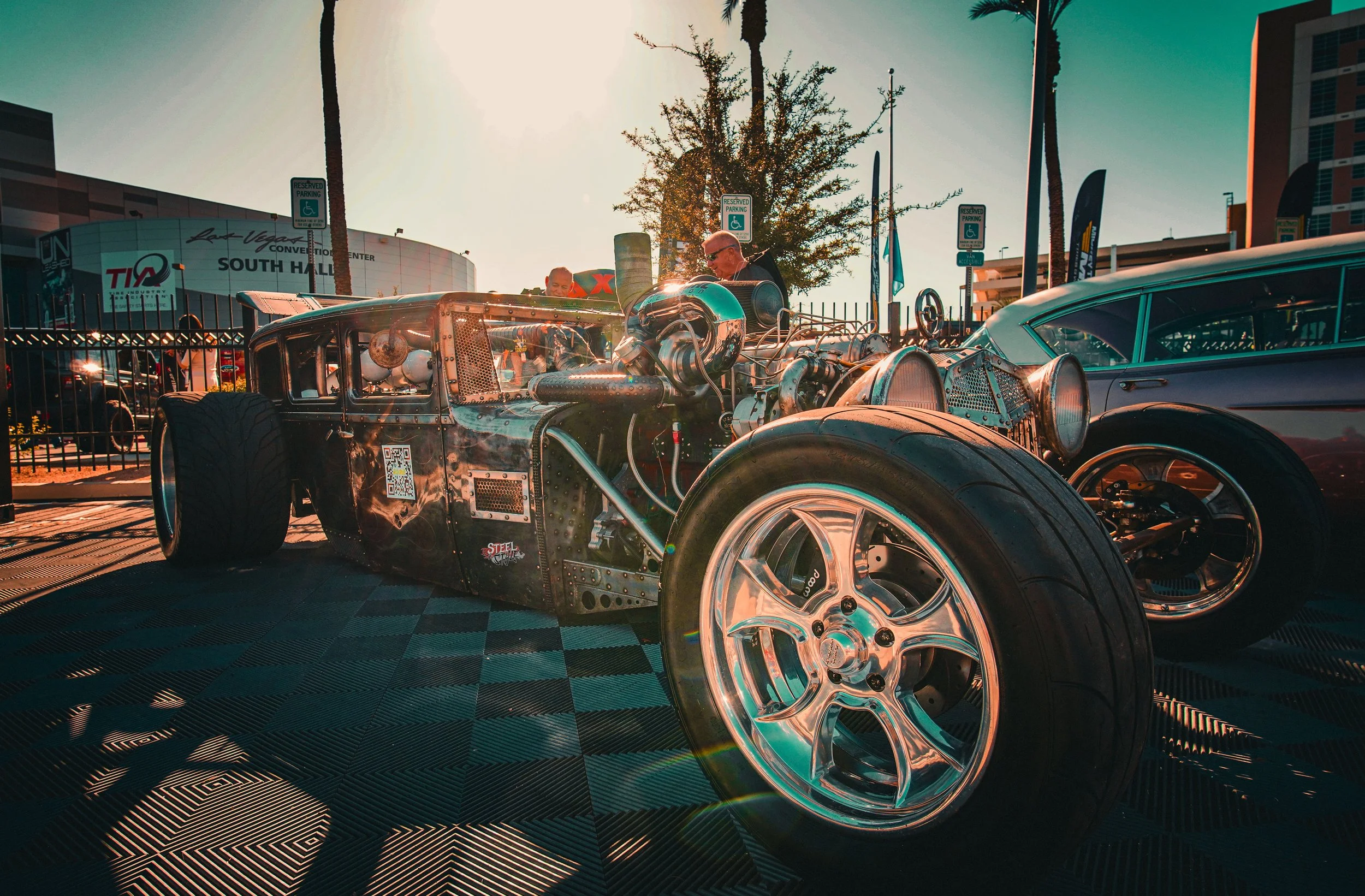 A vintage drag racing car on display outdoors with a clear sunny sky and palm trees in the background. Two men are inspecting the car, which has an exposed engine and shiny chrome wheels. Behind them, there are signs indicating reserved parking space