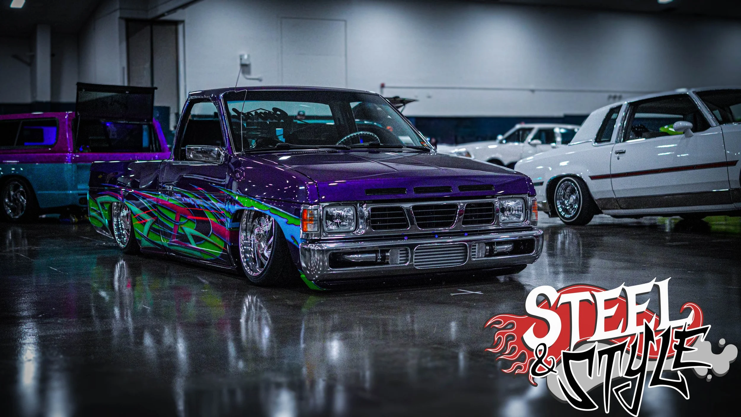 A purple customized pickup truck with vibrant green and blue flame graphics parked indoors at a car show, with other vintage cars visible in the background.