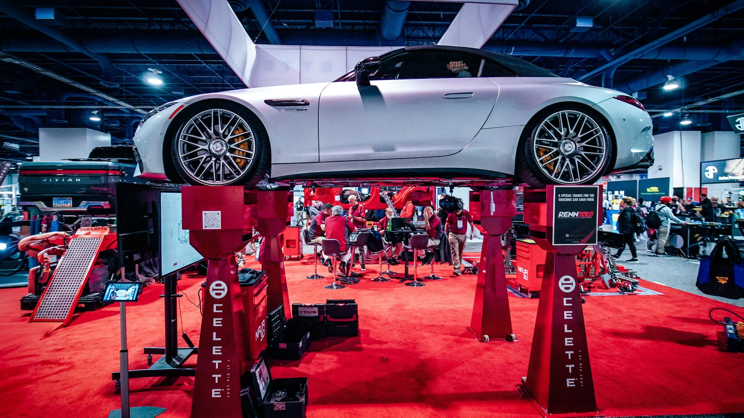 A white sports car elevated on red stands at an indoor auto show, with people and booths visible in the background.