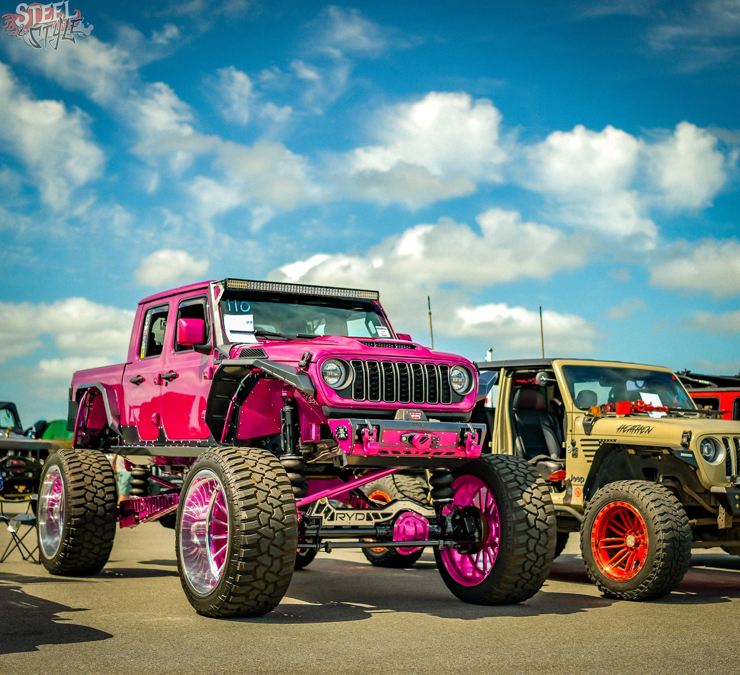 A pink lifted Jeep with large off-road tires parked in a parking lot during the daytime, with a blue sky and scattered clouds above.