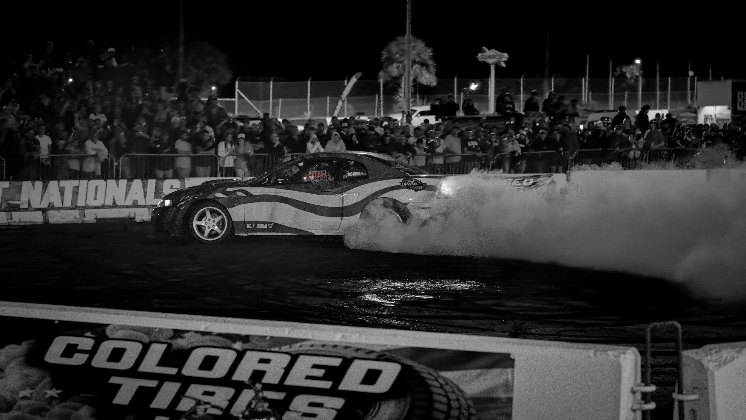 A race car performing a burnout on a track at night, with smoke coming from the rear tires, and a crowd of spectators standing behind a fence watching.