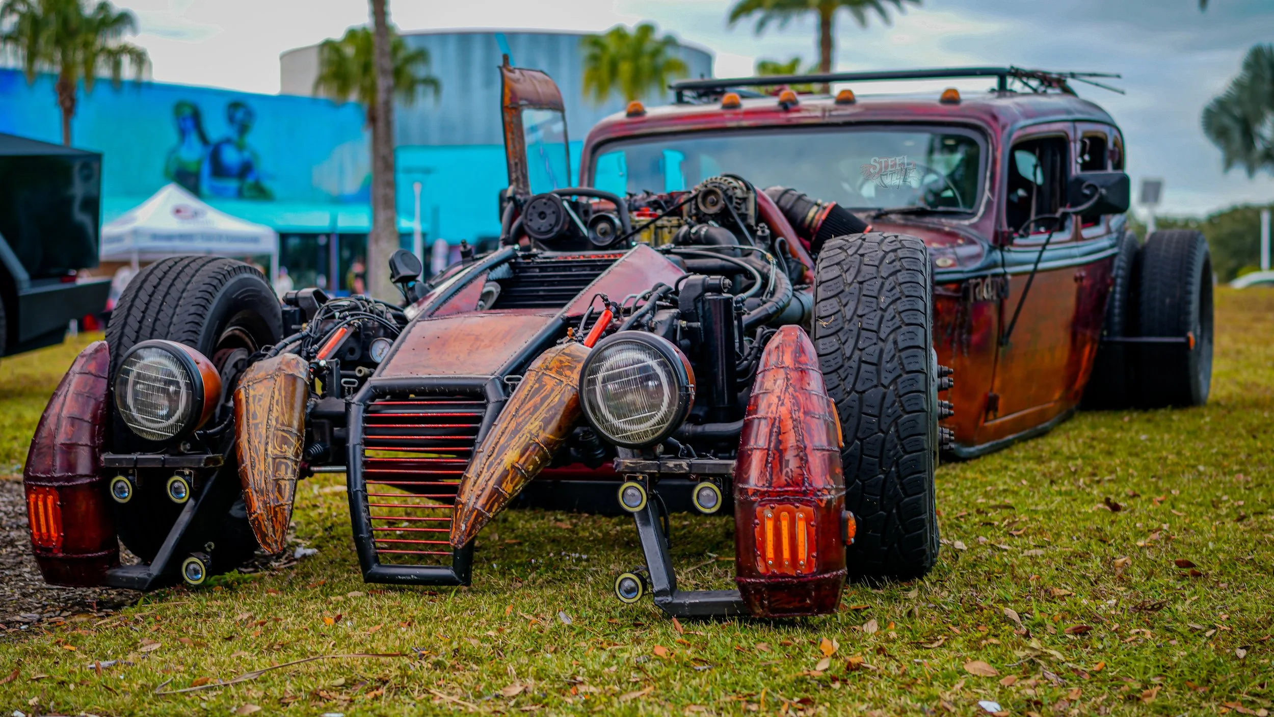 A custom-built, modified car with exposed engine components, wide tires, and a vintage style, displayed on grass with trees and tents in the background.