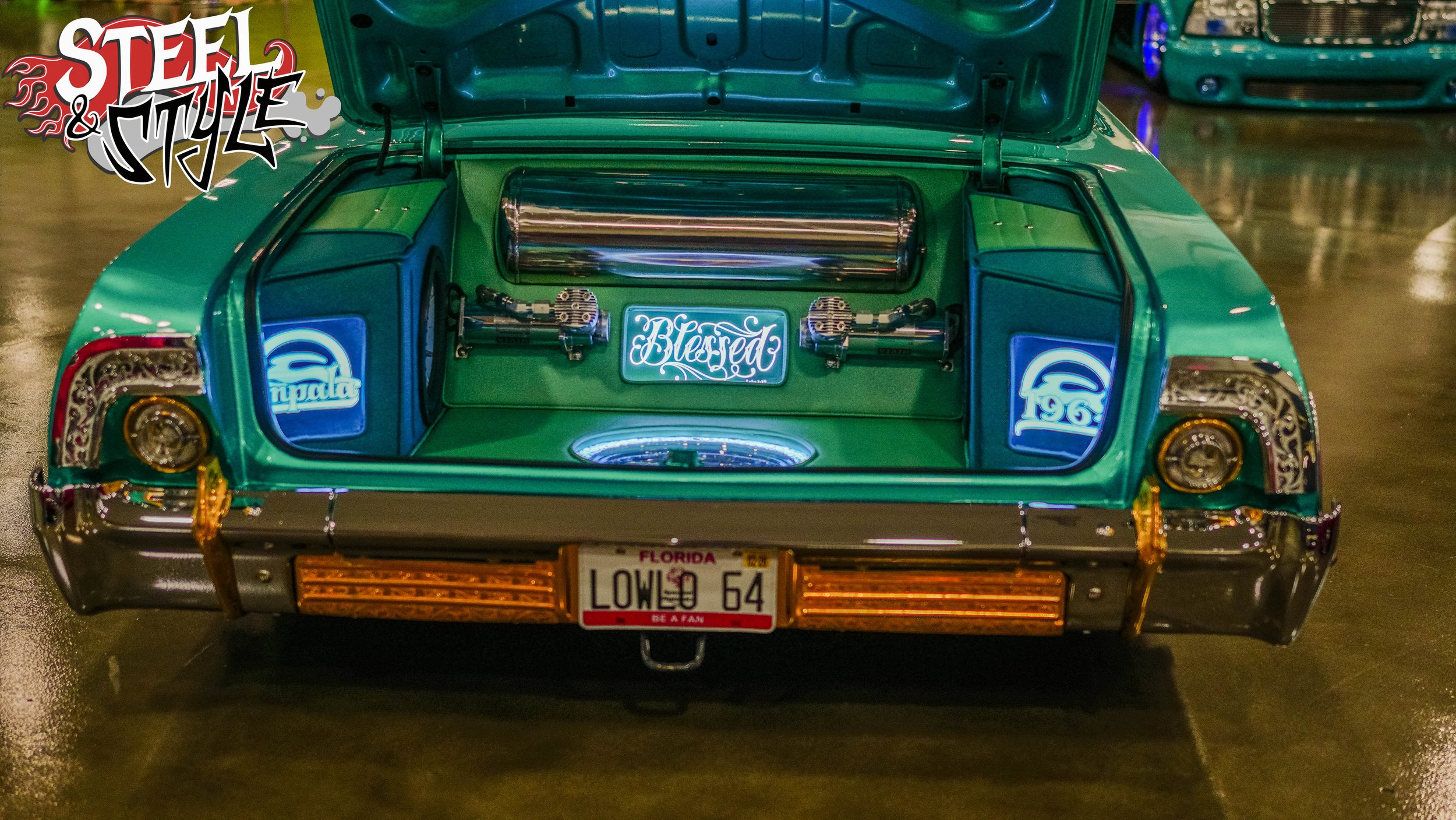Rear view of a vintage car displayed indoors with its trunk open, revealing custom interior with neon signs reading 'Blessed' and 'Impala,' a metallic tank, and car parts. The car has a Florida license plate that reads 'LOWED 64'.