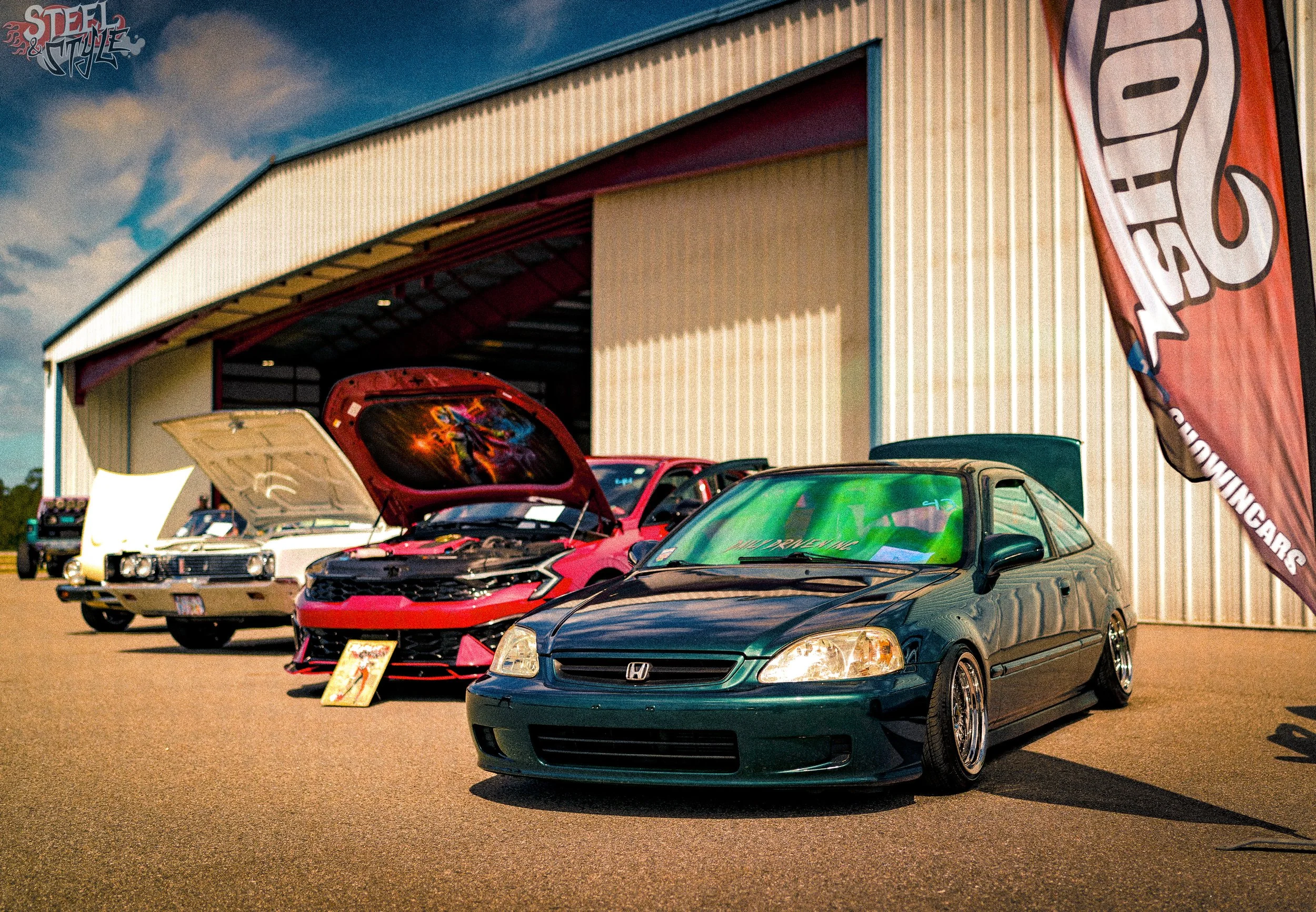 A lineup of modified cars displayed outside a building with a banner that reads 'Speed'. The cars have custom paint jobs and aftermarket parts, and are parked on a paved lot under a partly cloudy sky.