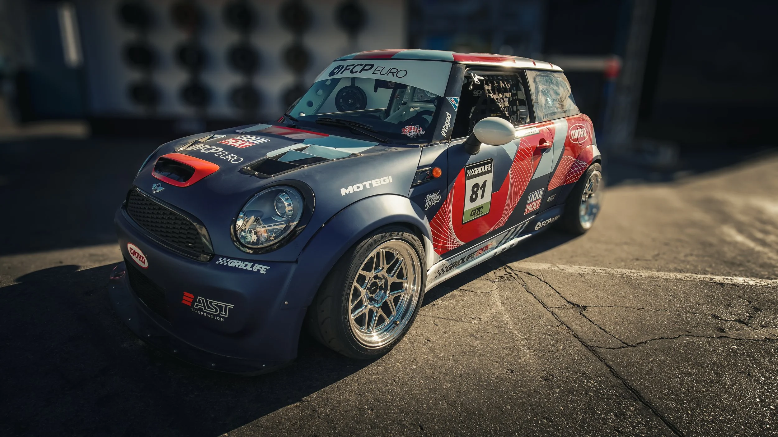 A race car with a blue body and red and white accents, parked on asphalt, featuring various sponsor stickers including 'FCP Euro,' 'Motegi,' 'GridLife,' and 'Liqui Moly,' with racing number 81 on the door, and equipped with aftermarket wheels and a m