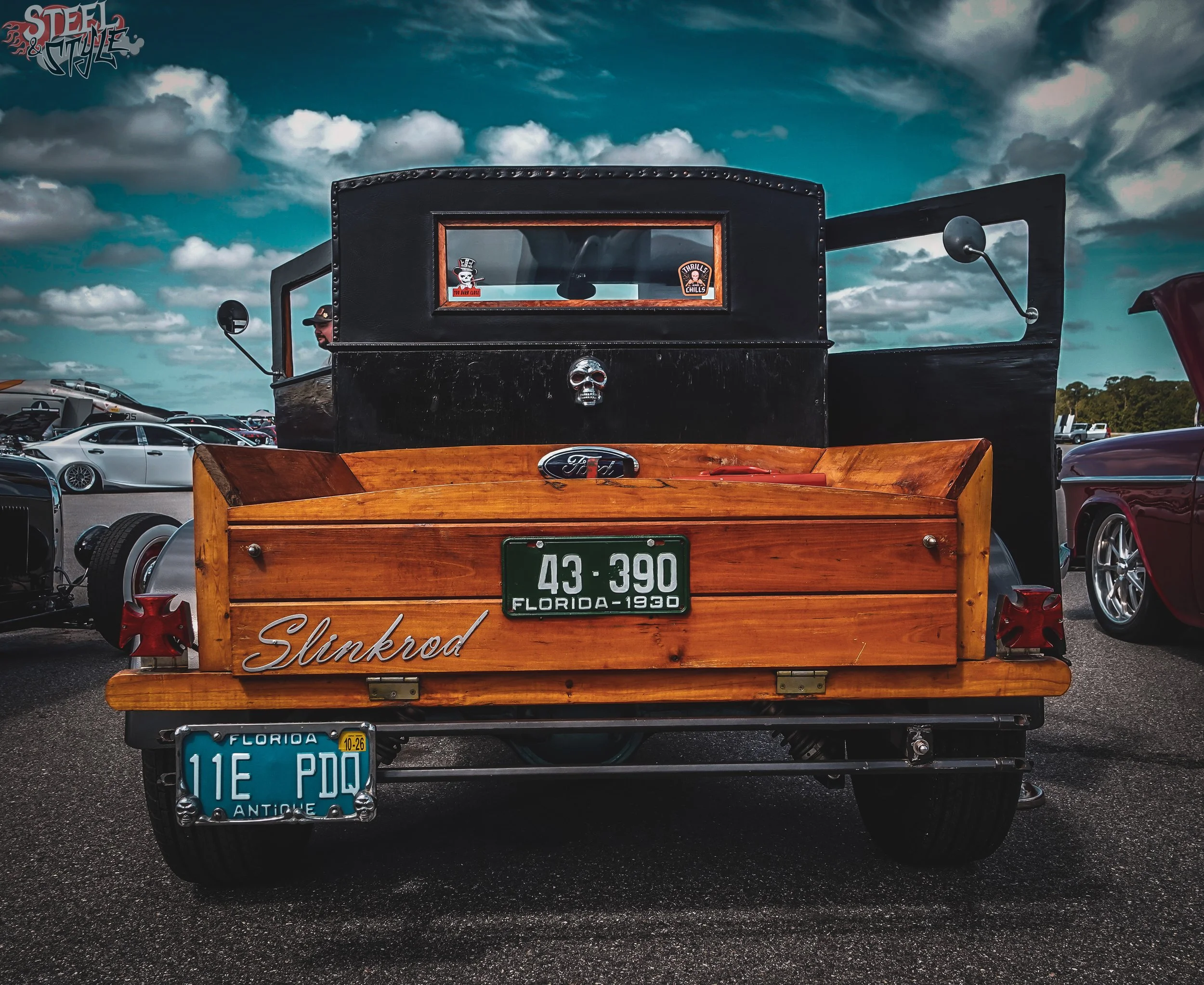 Back of a vintage wooden cart with a black top and Boyd Coddington license plates, parked in a lot filled with modern cars and a cloudy sky overhead.