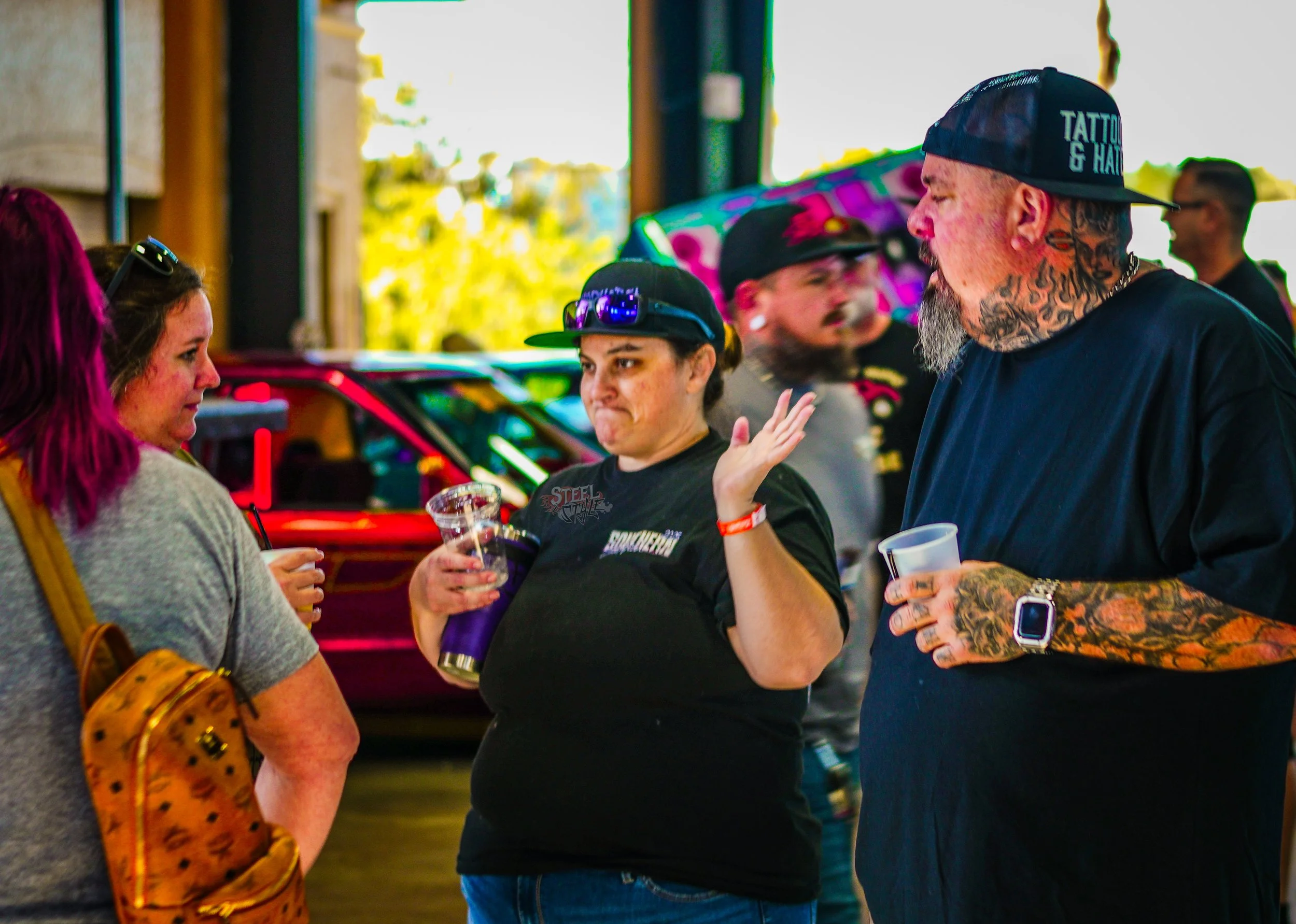 Group of people conversing at an outdoor event, with colorful cars in the background, some holding drinks and wearing casual attire.