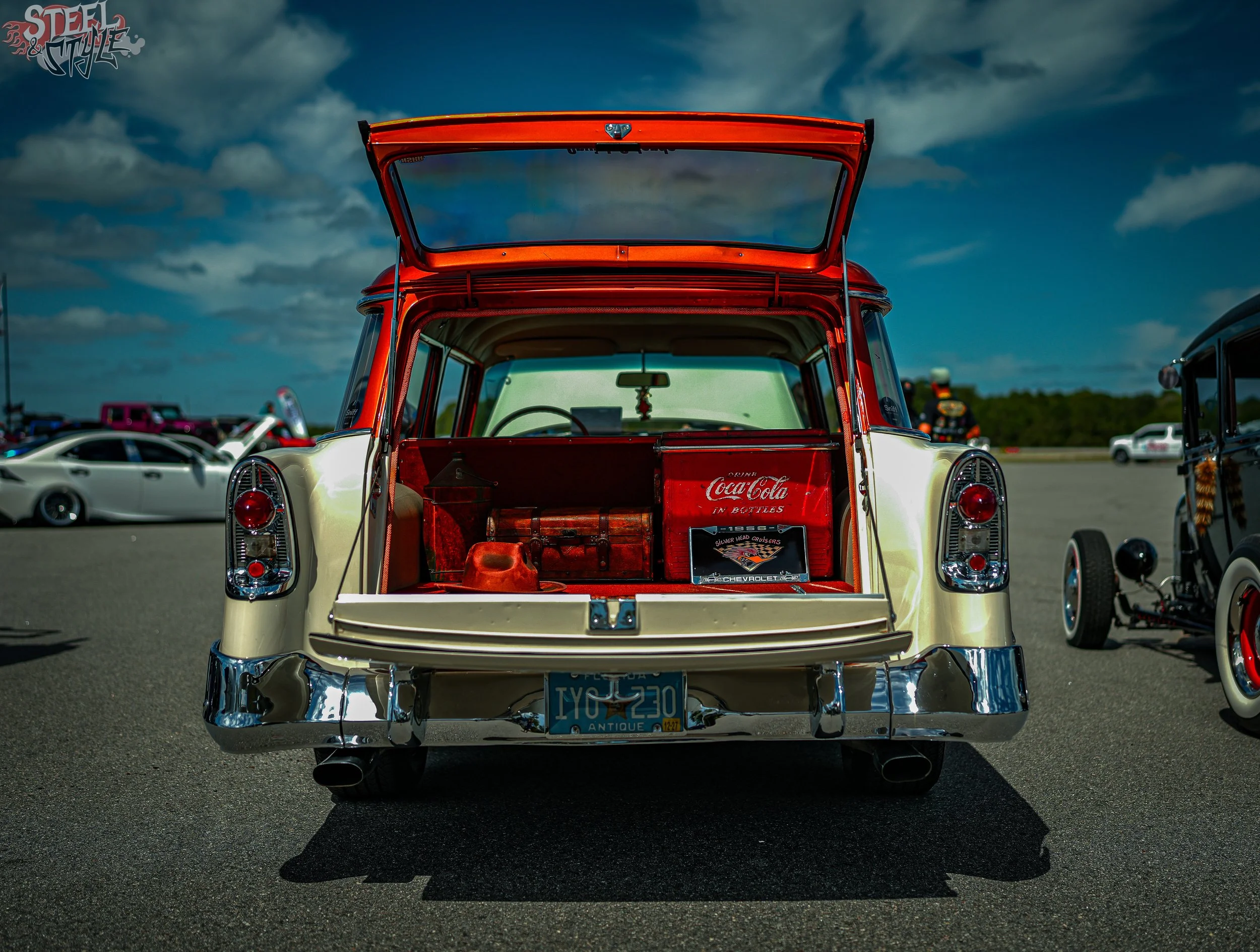 The image features a vintage Chevrolet car with its rear hatch open at a car show, displaying a Coca-Cola vending device and various car memorabilia inside. The car has a shiny chrome bumper, classic tail lights, and a license plate that reads "YCC 3