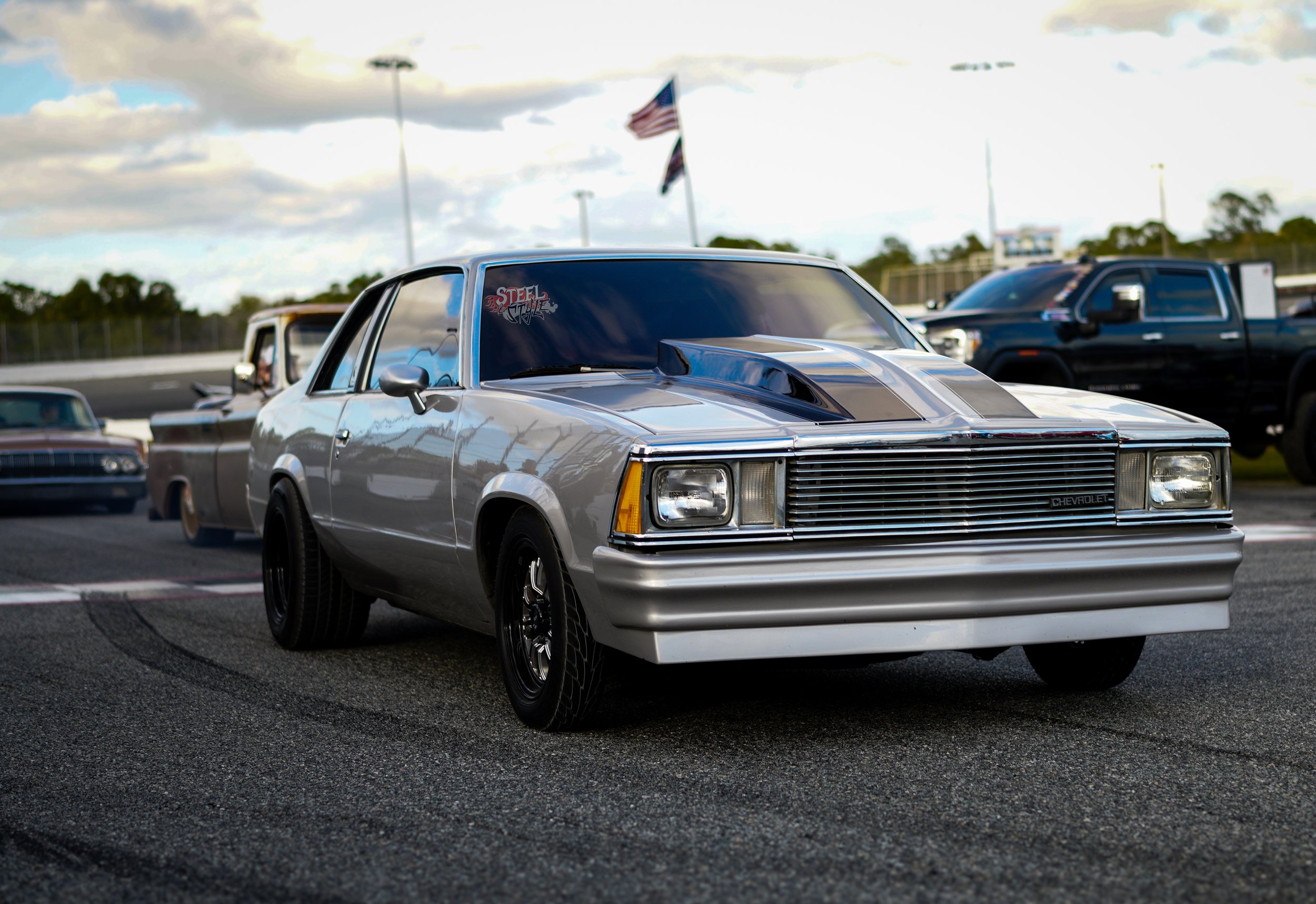 A vintage silver Chevrolet car with black wheels parked on a race track, with other classic cars and trucks in the background, American flags in the sky, and cloudy weather.