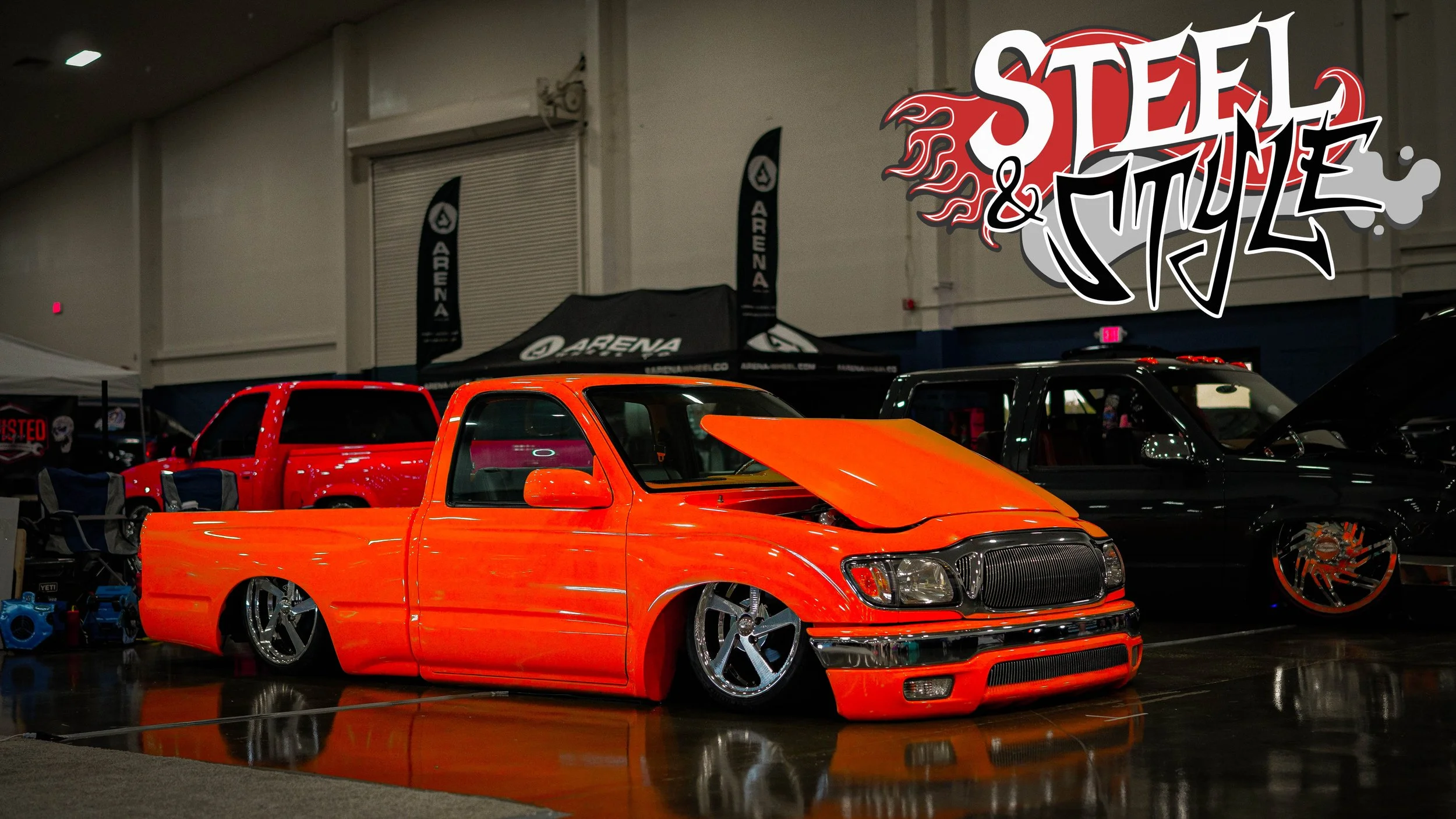 An orange customized pickup truck with a lowered stance and large chrome wheels, displayed at an indoor car exhibition with a black and red largest logo reading 'Steel & Style' in the top right corner.