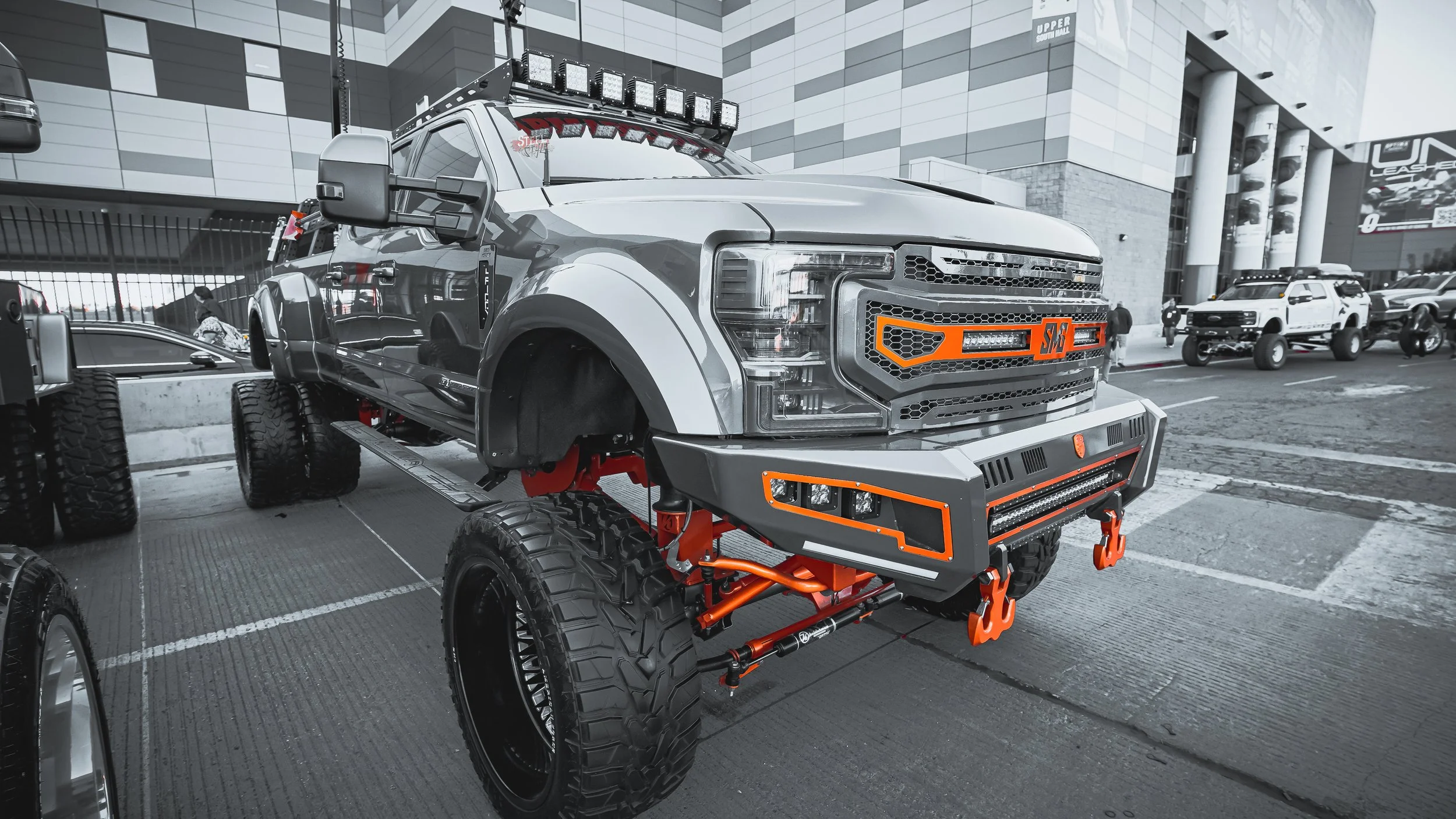 A heavily modified off-road truck with oversized tires, a custom front bumper, orange accents, and additional lighting, parked in an urban setting.