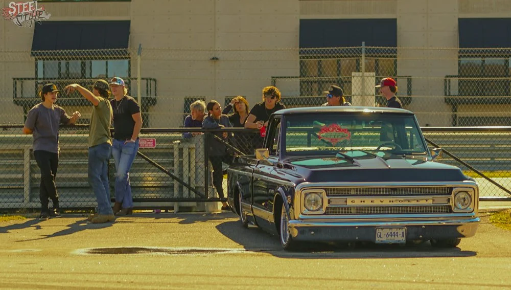 A group of people standing near a black vintage Chevrolet truck with a custom decal on the windshield. They are on a sidewalk next to a chain-link fence, with a building in the background.