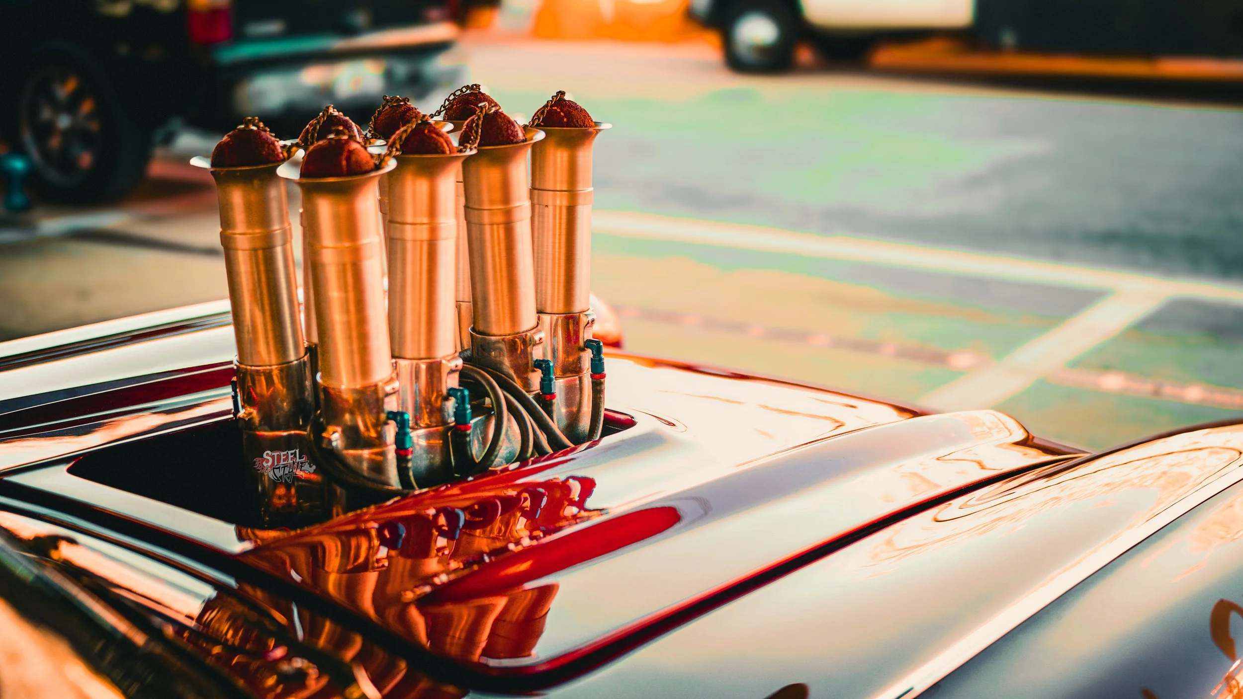 A car with a rocket engine mounted on its hood, with four large copper tubes shaped like turbojets and topped with red spherical objects, possibly model rockets.