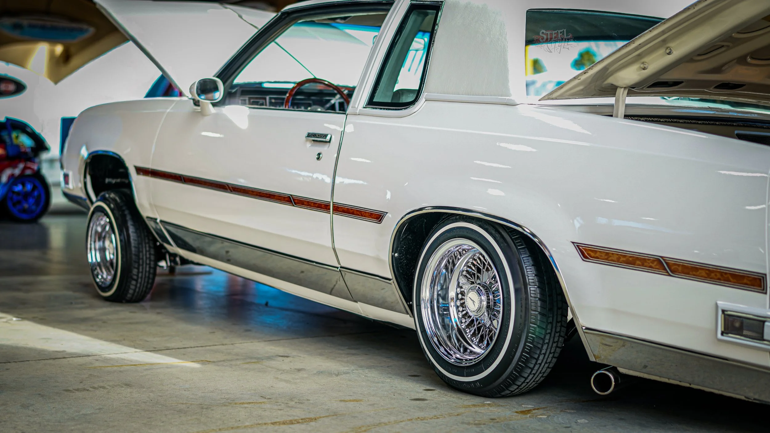 A white vintage car with chrome wire wheels parked indoors with the hood open.
