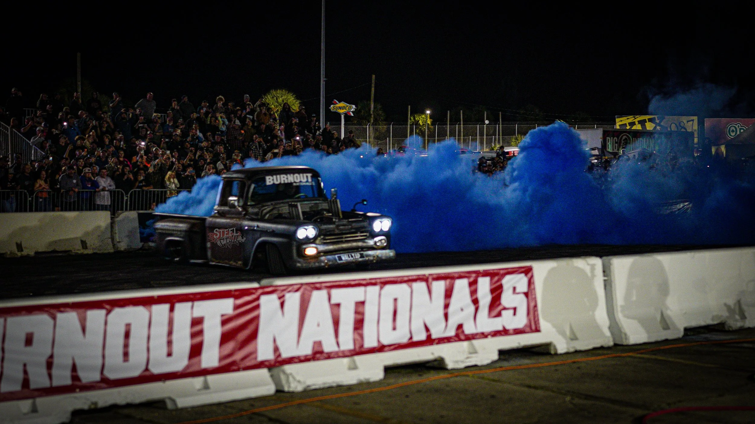 A vintage car, painted black with the word "BURNOUT" on the windshield, performs a burnout on a dirt track at night, creating blue smoke. The track is lined with concrete barriers and a banner reading "BURNT NATIONALS." In the background, a large cro
