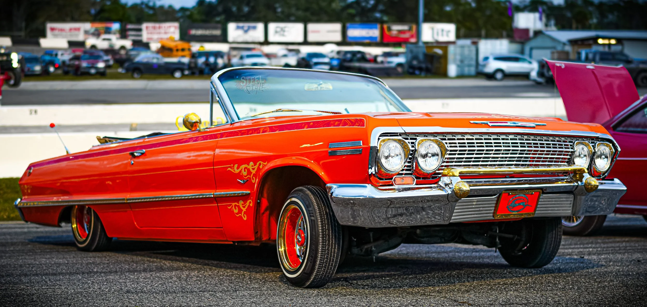 A vintage red convertible car with gold accents and decorative flame decals parked at a car show. The car has a chrome grille and bumper, and the hood is open. In the background, there are other cars and a parking lot with billboards and trees.